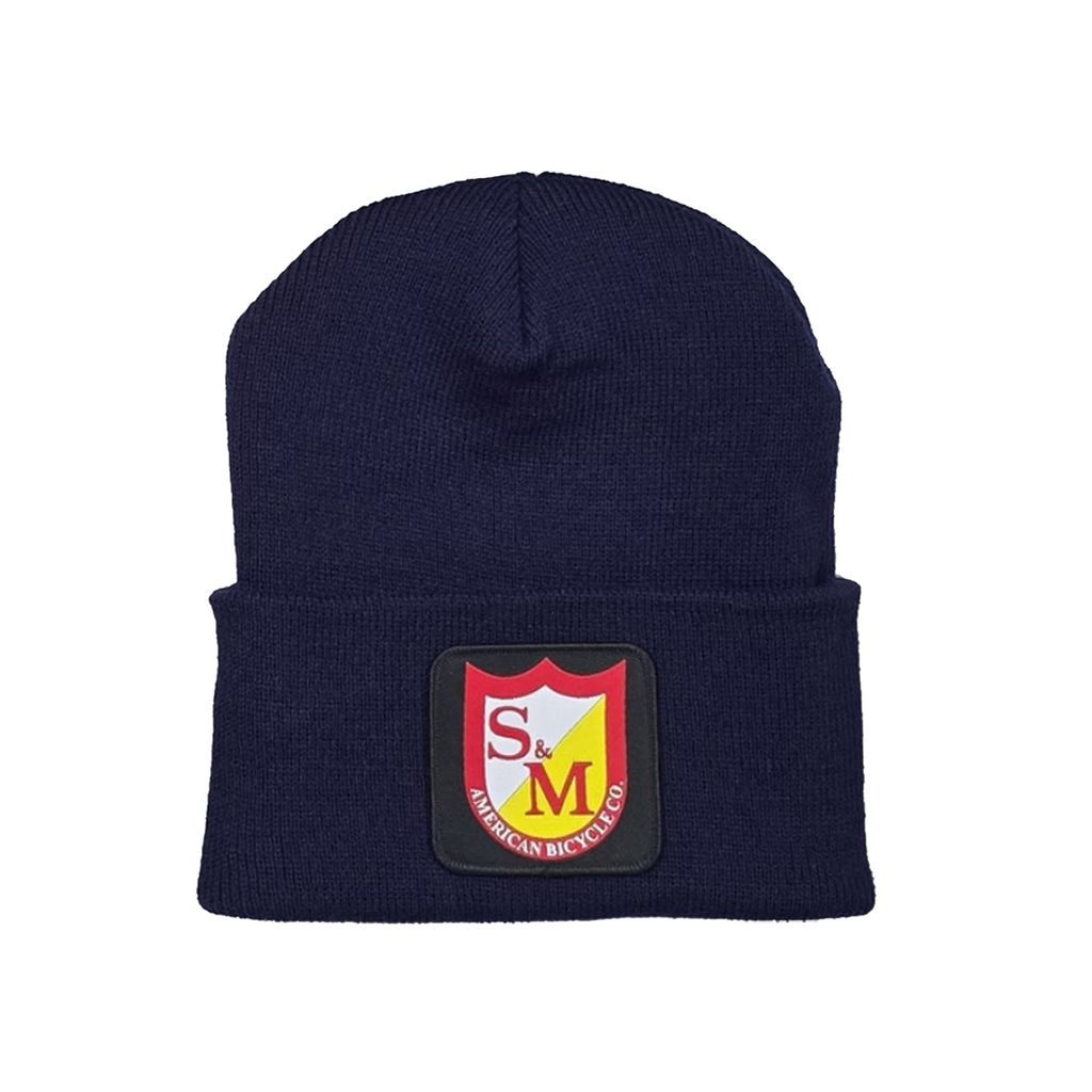 The S&M Dense Cuffed Beanie, in navy blue and made from hypoallergenic acrylic, is adorned with a red and yellow shield-shaped patch labeled "S&M American Bicycle Co." It provides a comfortable fit, accommodating most head sizes.