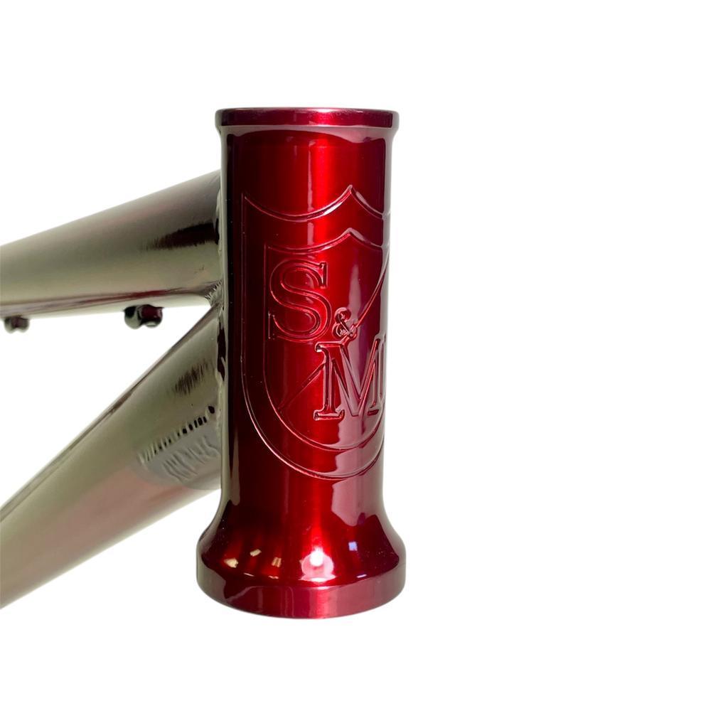 Close-up of the S&M Big Jumper V2 26 Inch Dirt Jumper Frame's metallic red head tube, featuring an embossed S&M logo and shield, built for suspension forks and disc brake compatibility.