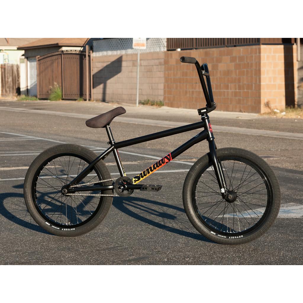 A Sunday Sound Wave Special 20 Inch Bike (2023) with thick tires, Odyssey parts, and a brown seat stands upright on an asphalt surface in a residential area.