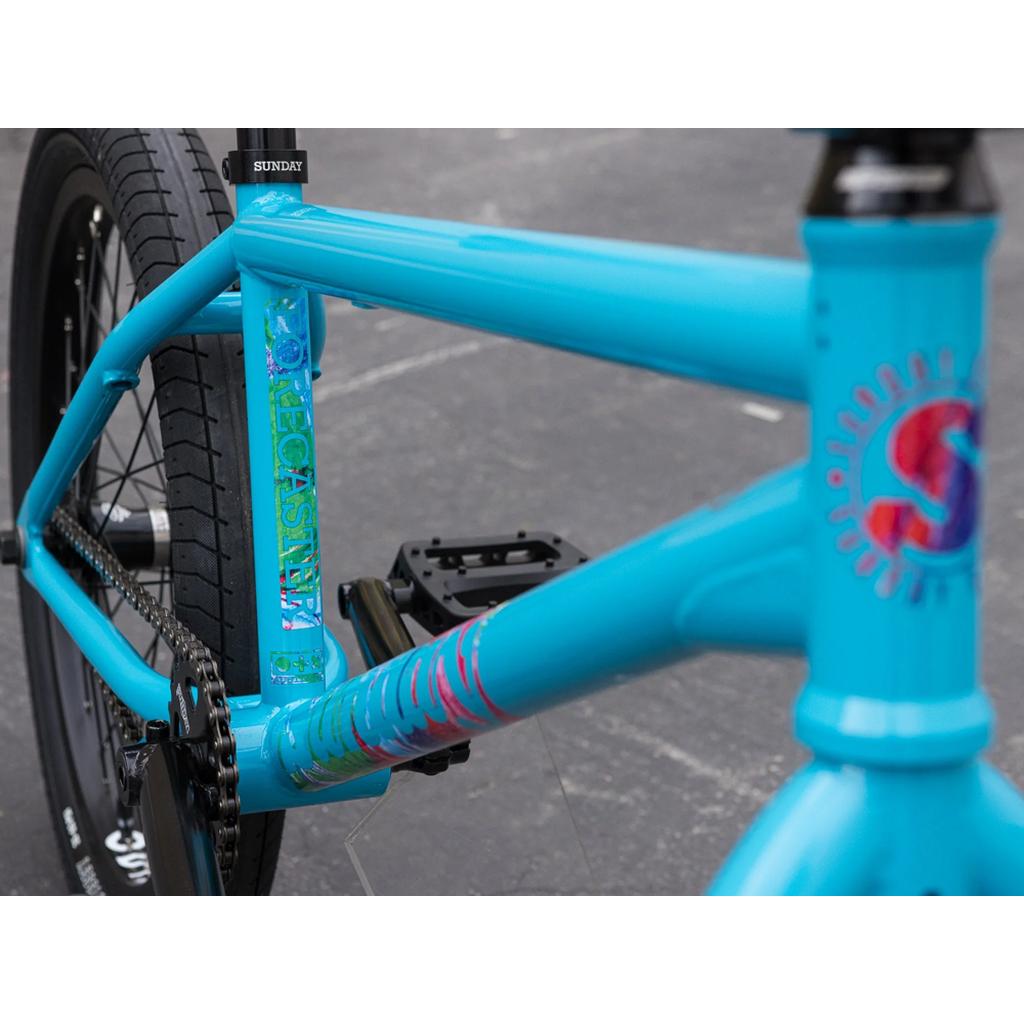 Close-up of a Sunday Forecaster 20 Inch Bike (2025) / Aaron Ross in blue, featuring colorful decals, Odyssey components, black pedals, and black tires on a concrete surface.