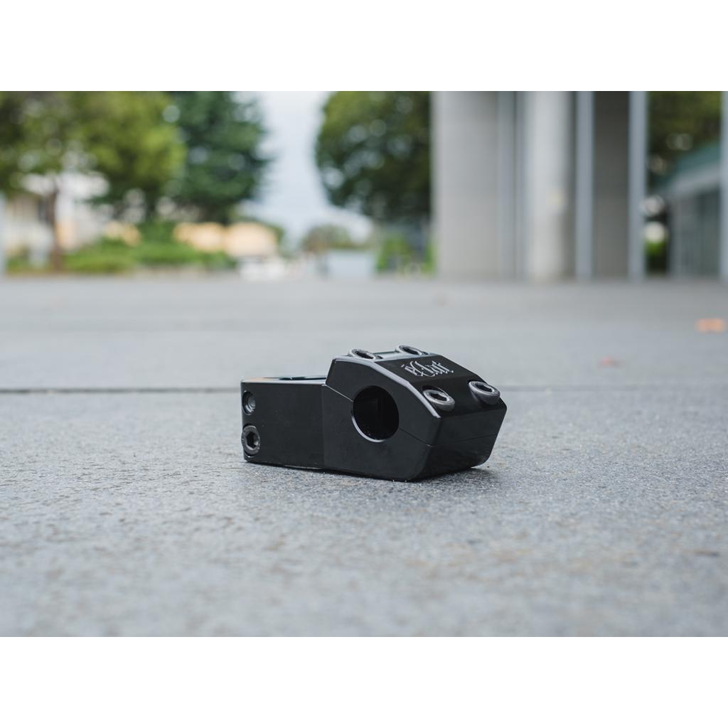 The Eclat 1023 Stem, a limited-edition black BMX stem with unique mid-school styling, lies on a concrete surface with blurred trees and buildings in the background.