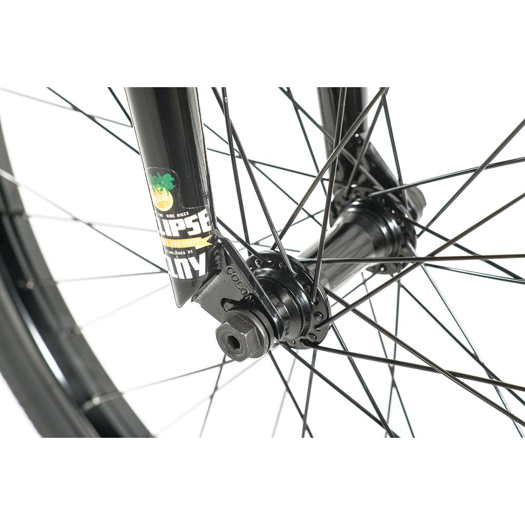 Close-up of the Colony Eclipse 24 Inch Bike’s front wheel, featuring black spokes, hub, axle, fork with a colorful sticker, and chromoly frame details—a stylish BMX cruiser set against a white background.