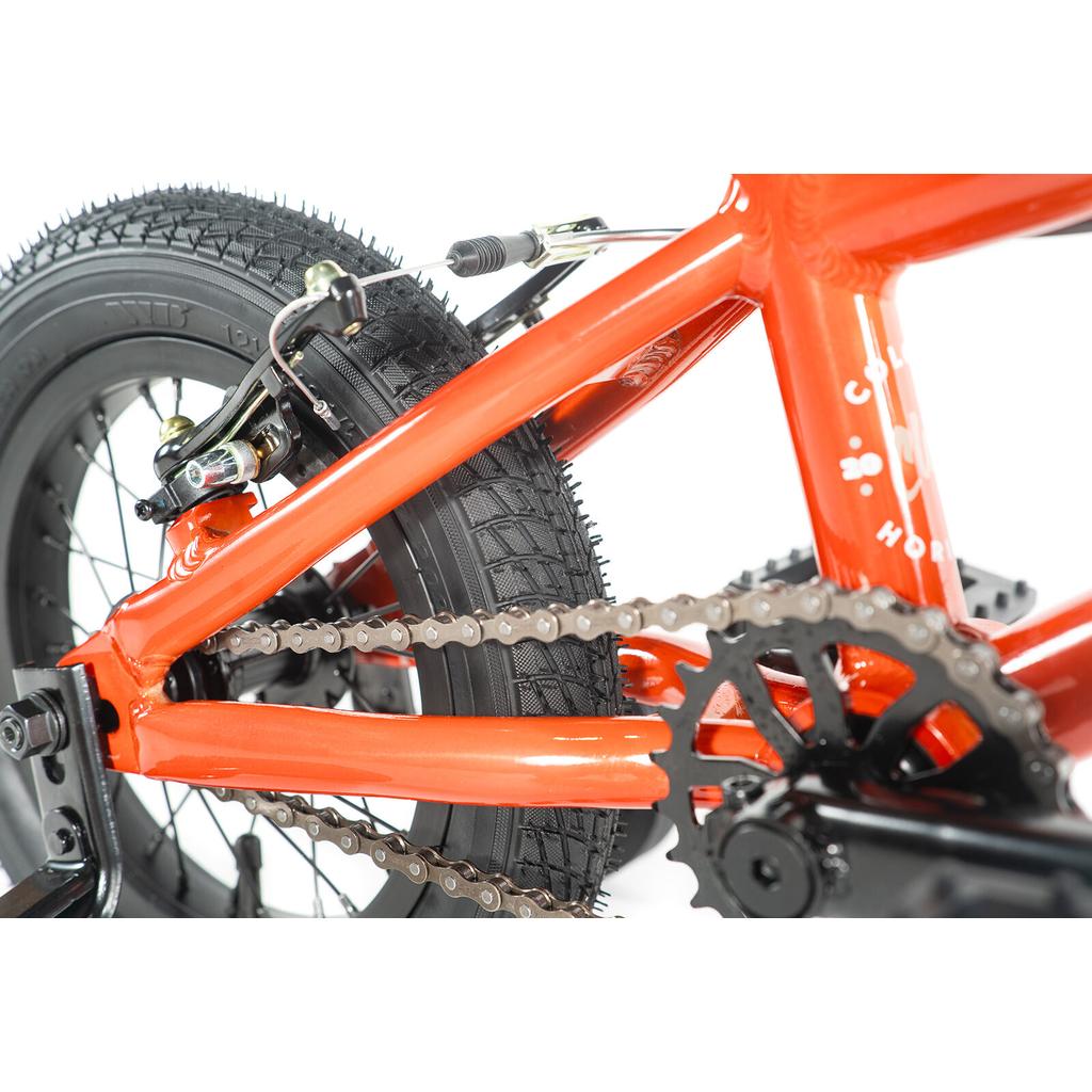 Close-up of the rear wheel, chain, gears, and brake mechanism on an orange Colony Horizon 12 Inch Bike with a sturdy alloy frame.