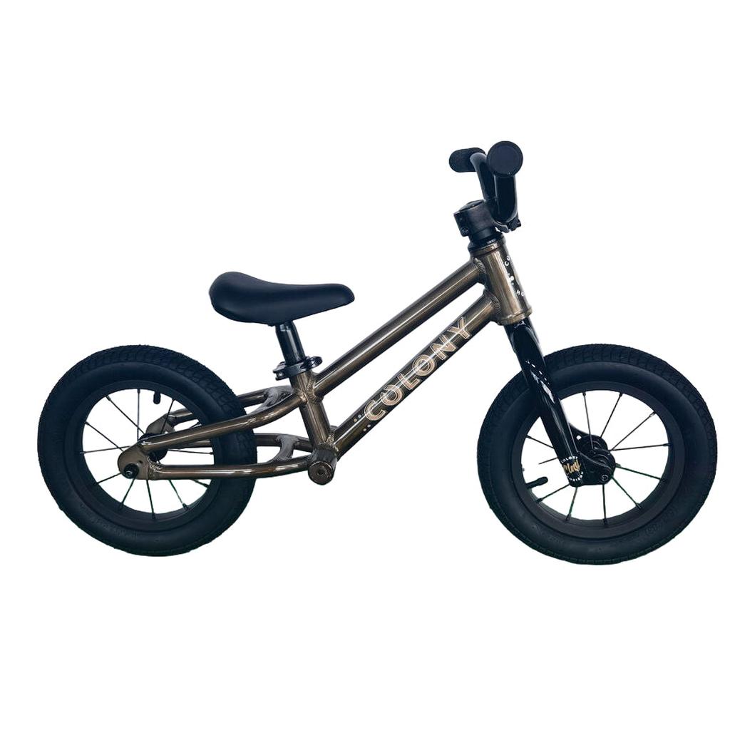 A Colony BMX Horizon Alloy Balance Bike with black tires and a dark metallic frame labeled "COLONY," placed on a green floor against a white wall.
