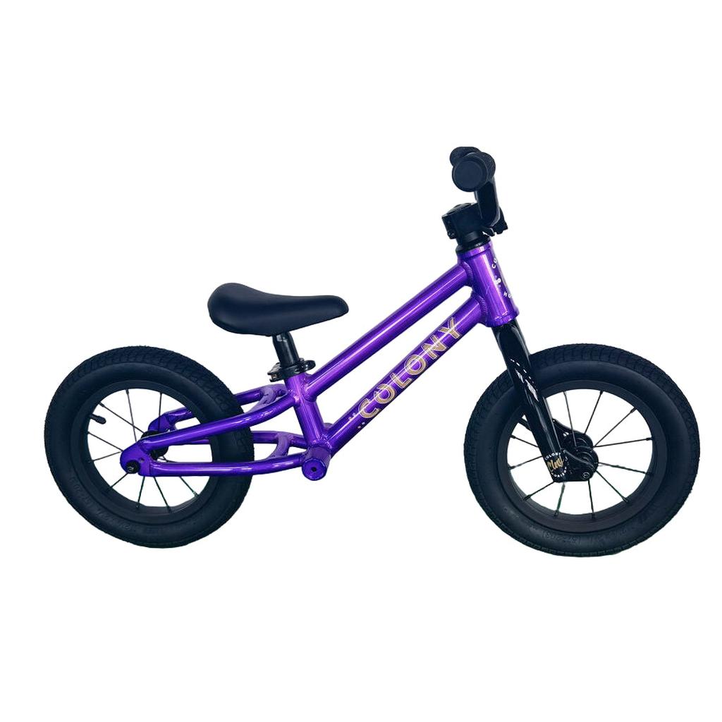 The Colony BMX Horizon Alloy Balance Bike, a small purple kids' bike with black wheels and handlebars, is displayed on a green floor against a white wall.