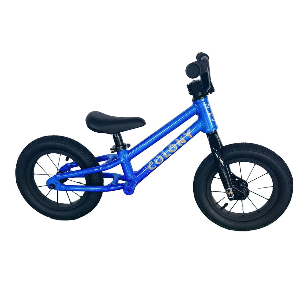A small blue Colony BMX Horizon Alloy Balance Bike for kids, featuring black tires and handlebars, sits on a green floor against a white wall. Ideal as a first BMX balance bike for young riders.