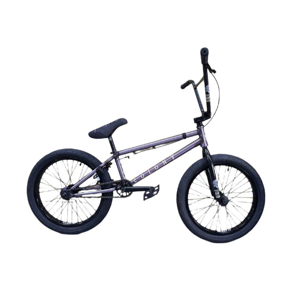 A Colony Rico Elite 20 Inch Bike in purple with black tires and Guardian forks is parked in front of stacked cardboard boxes in a warehouse.