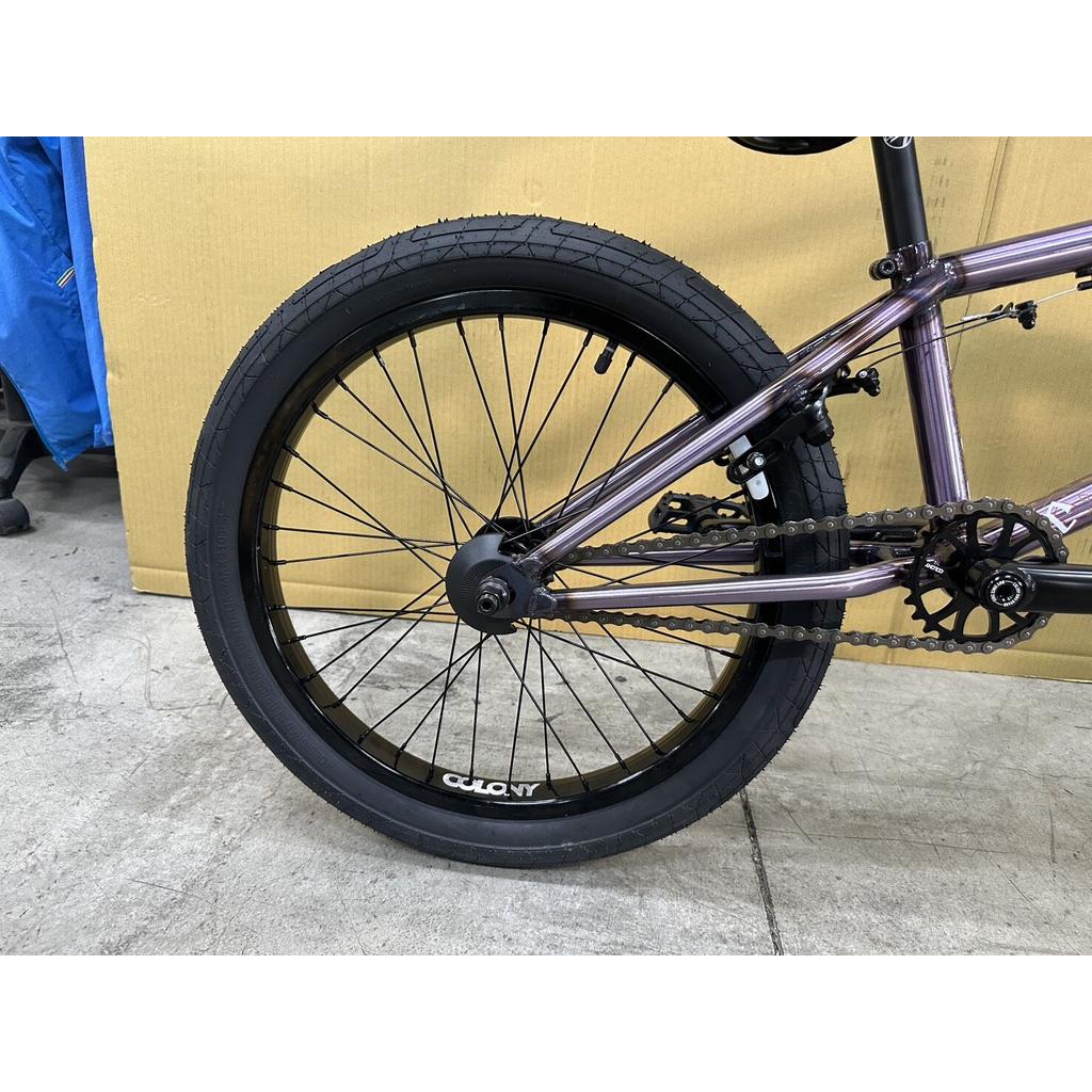 Close-up of the rear wheel and gear mechanism of the Colony Rico Elite 20 Inch Bike, featuring a durable CrMo frame, set on a concrete floor against a brown background.