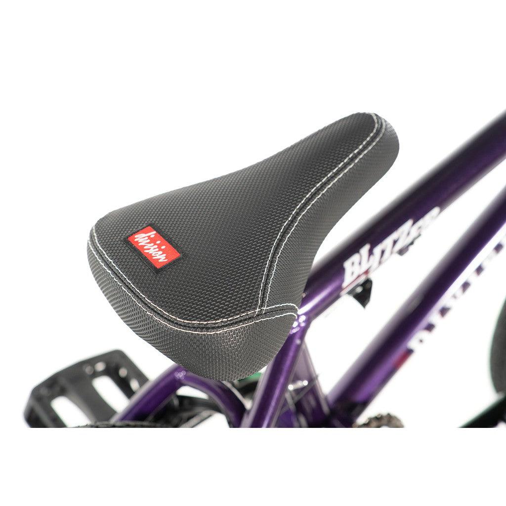 Close-up of a bicycle seat featuring a textured black cover and white stitching on the purple frame of an entry-level BMX bike. A red label with white text is visible on the seat, highlighting the scaled-down geometry of the Division Blitzer 16 Inch Bike designed for young riders.