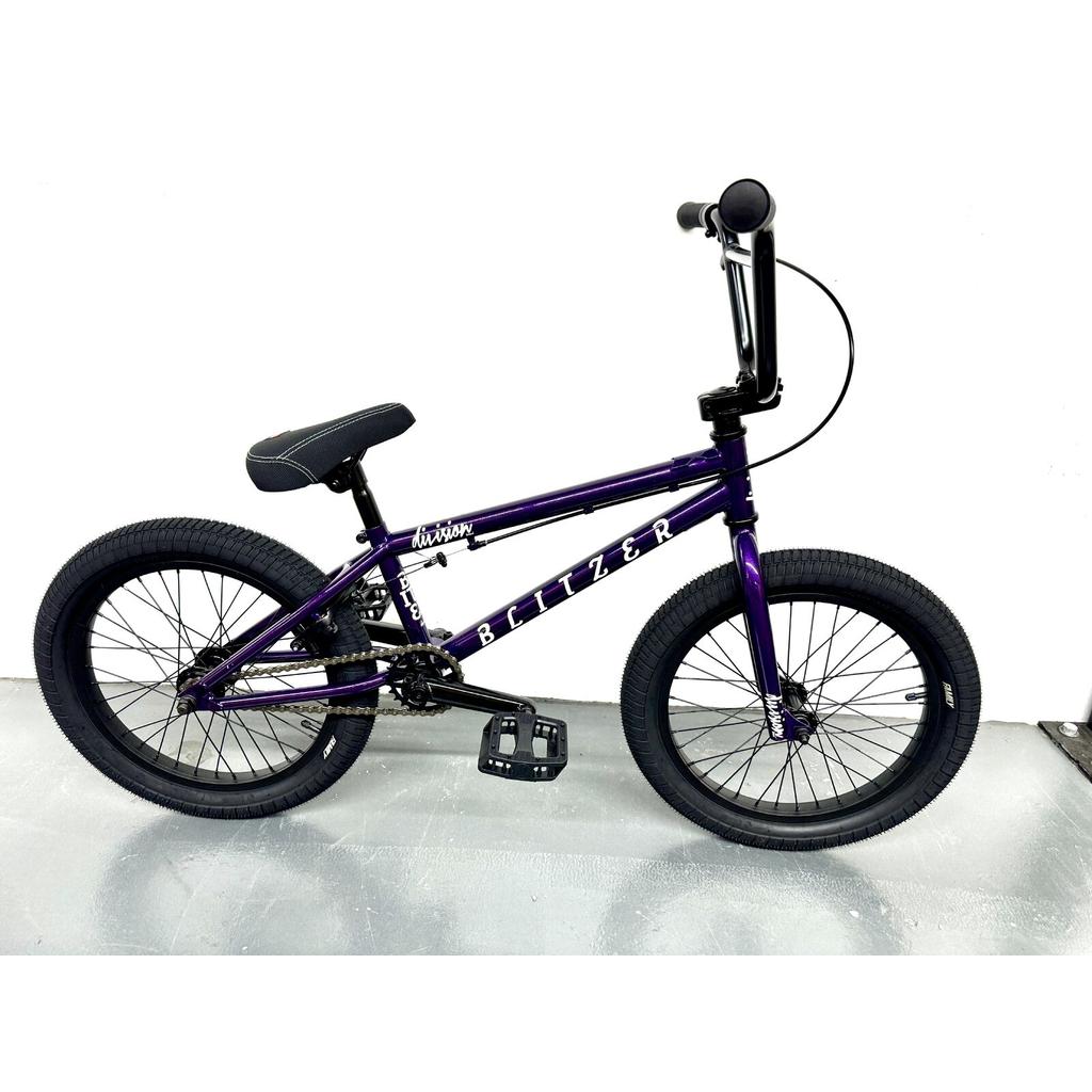 The Division Blitzer 20 Inch Bike, a purple beginner BMX with 20” wheels, black tires, and a black seat, stands against a plain white wall and gray floor.