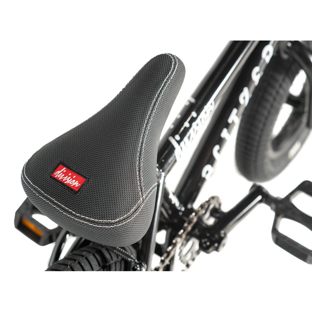Close-up of the Division Blitzer 12 Inch Bike's black kids BMX seat with white stitching and red logo on a black frame. Pedals and rear tire are partially visible.