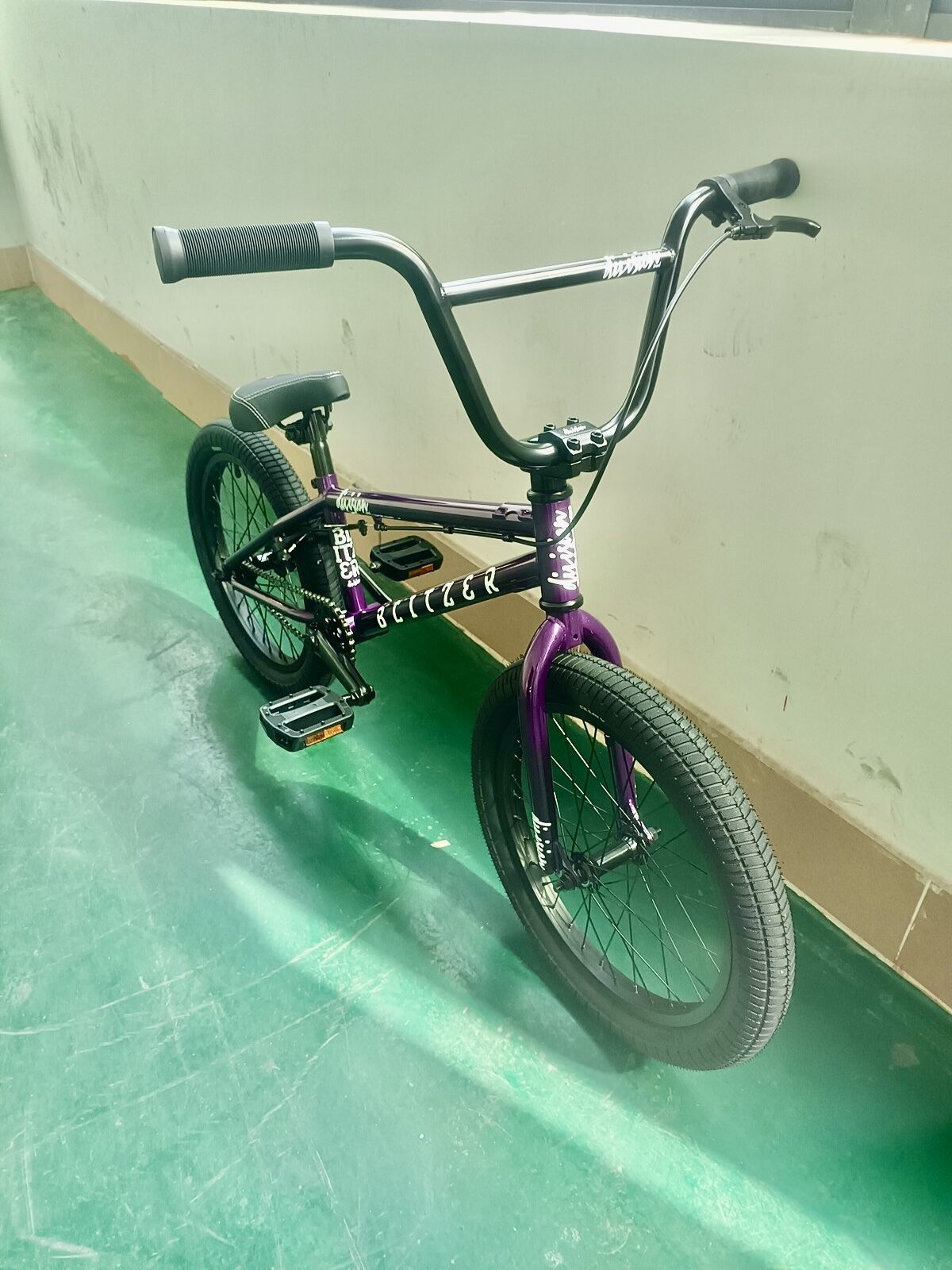 A Division Blitzer 18 Inch Bike in black and purple with a Hi-Ten frame is parked indoors on a green floor, leaning against a white wall.