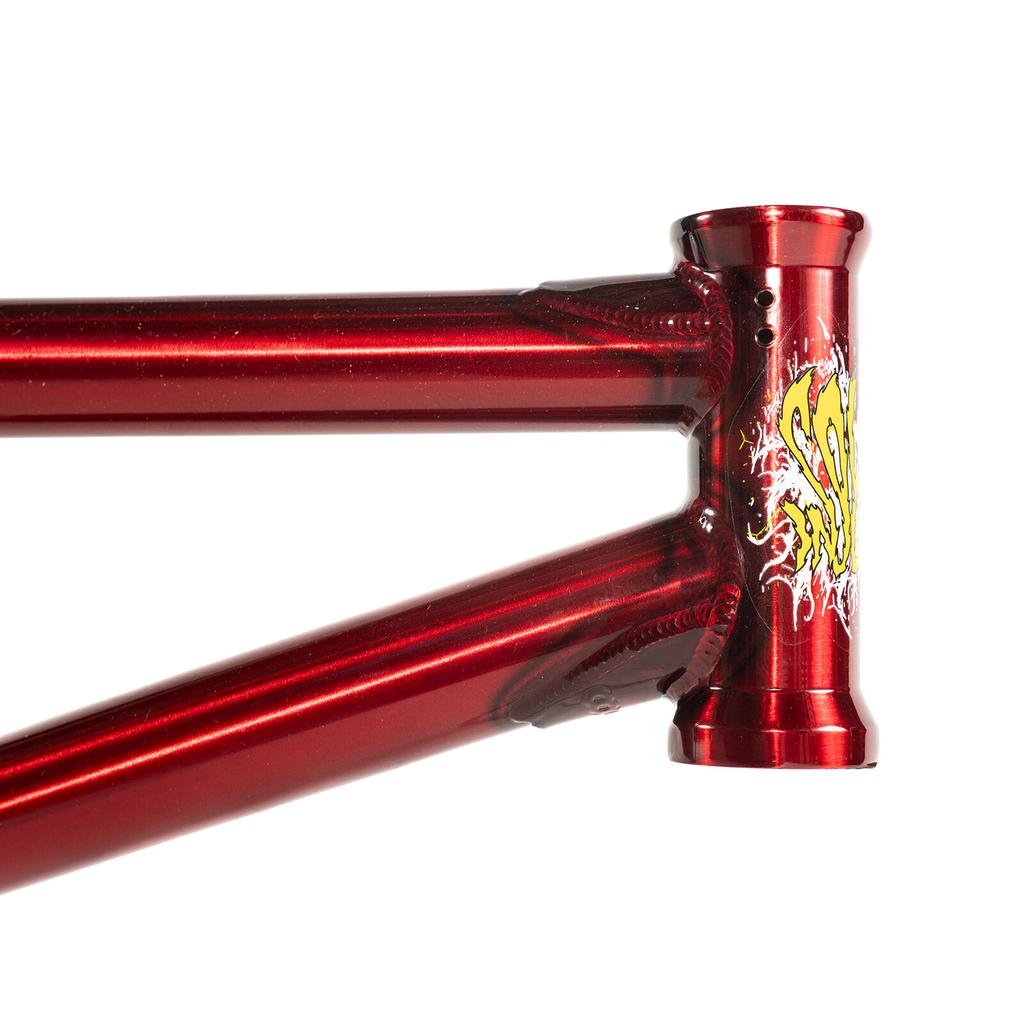 Close-up of the Colony Injustice Frame (Tom Justice Signature) in red metallic, highlighting the welds and head tube area, with a yellow and white graphic decal on the front. Crafted from 4130 CrMo for strength and durability.
