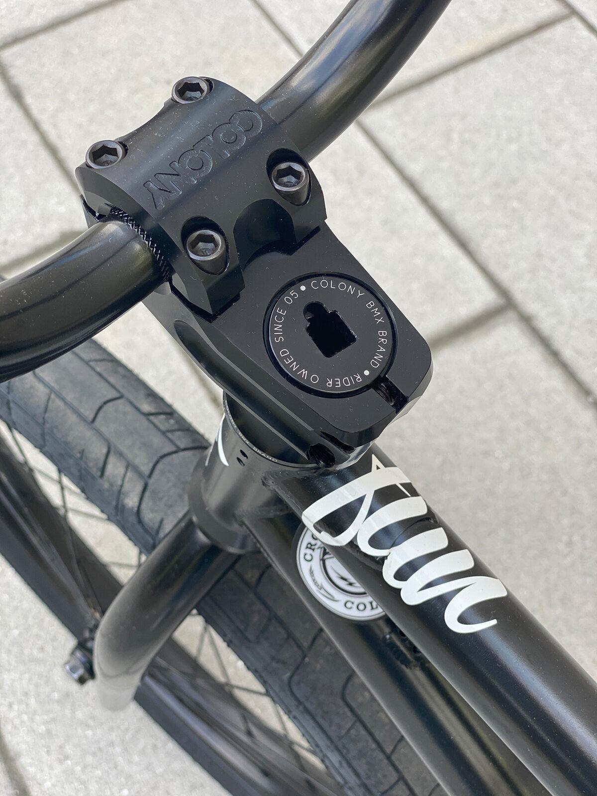 Close-up of a BMX bike handlebar with the Colony Squareback Stem in top load design, displaying the Colony logo and part of the frame with a white decal, set against a tiled pavement background.