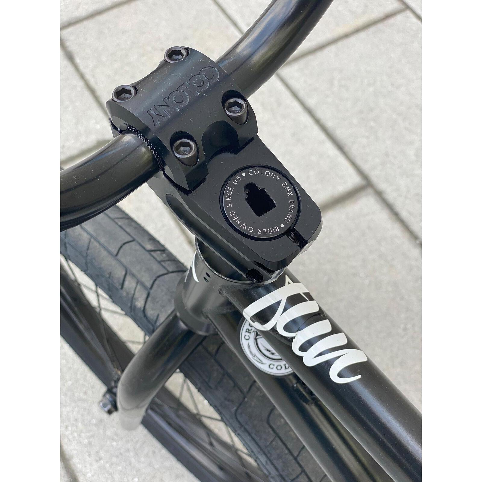 Close-up of a BMX bike handlebar with the Colony Squareback Stem in top load design, displaying the Colony logo and part of the frame with a white decal, set against a tiled pavement background.