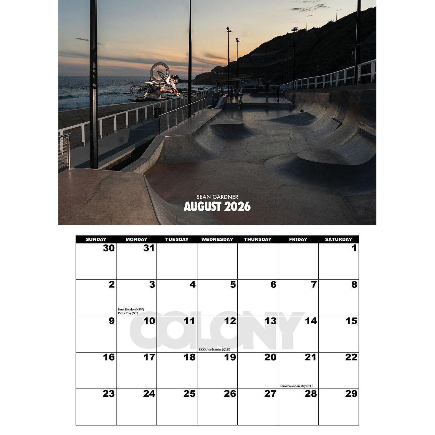 A BMX rider does a backflip above a concrete skatepark by the ocean at sunset, featured in the Colony 2026 Calendar for August.