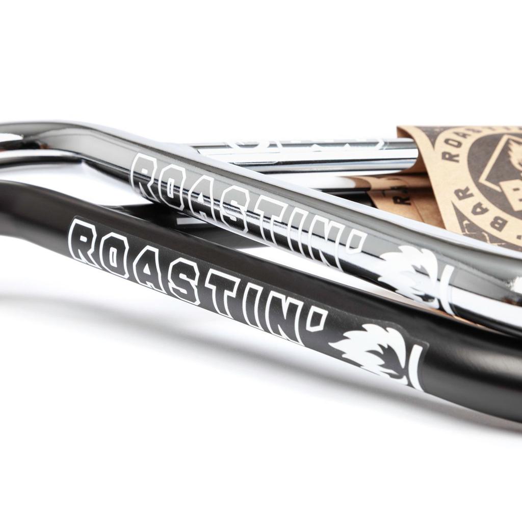 Close-up of two BSD Roastin Bars (Tam Roulston) chromoly handlebars—chrome and black—each with a "ROASTIN'" logo and white flame graphic, plus visible cardboard tags in the background.