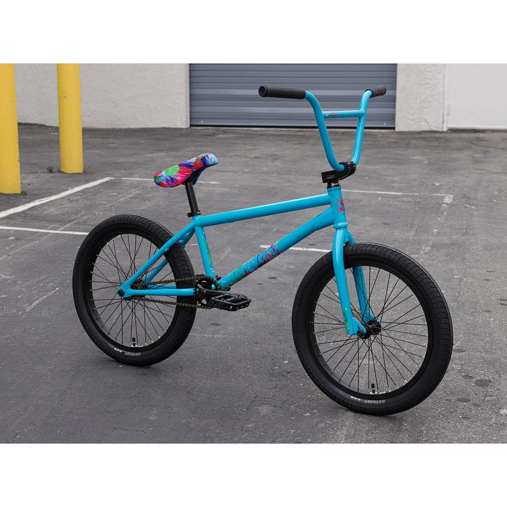 A Sunday Forecaster 20 Inch Bike (2025) / Aaron Ross, a blue chromoly BMX with a colorful seat and Odyssey components, is parked on gray pavement in front of a closed garage door and yellow poles.