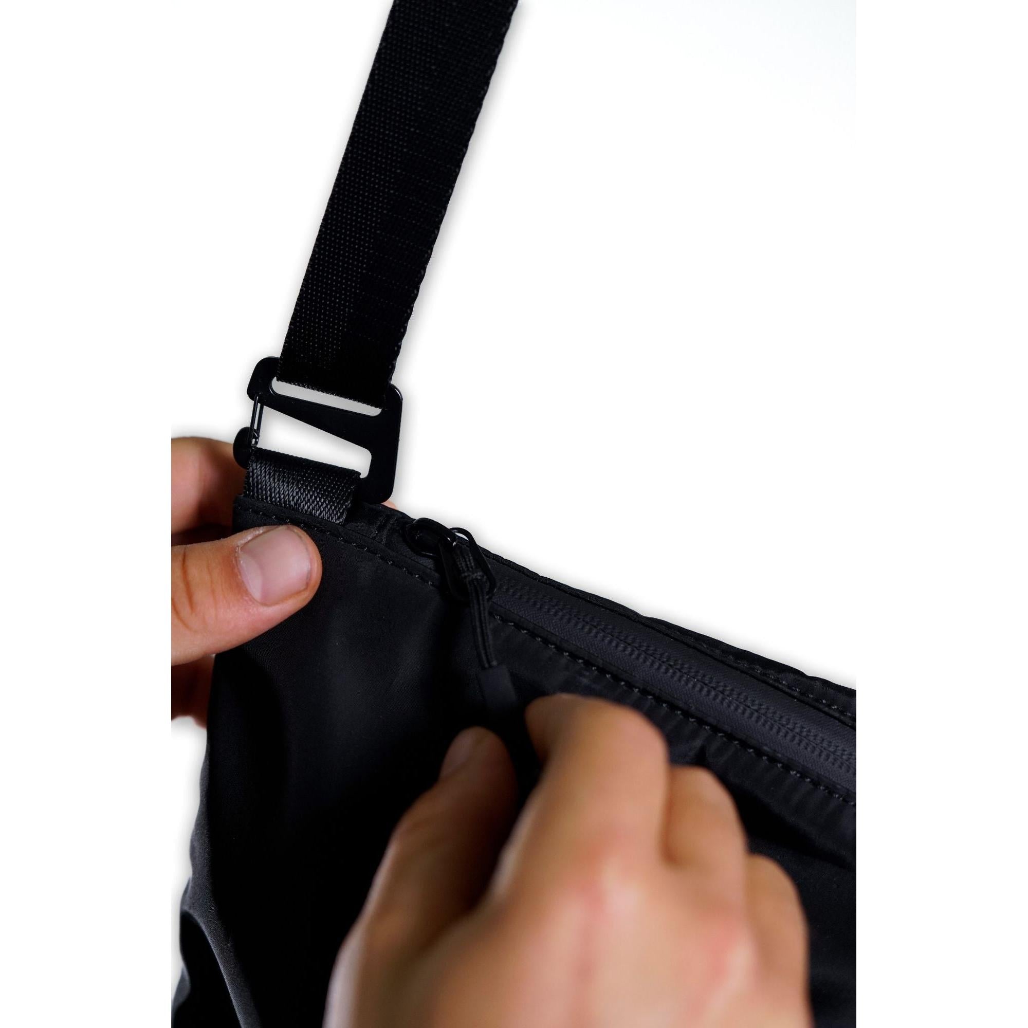 Close-up of hands adjusting a black strap and unzipping a black zipper on the V1da Everything Messenger Bag against a white background.