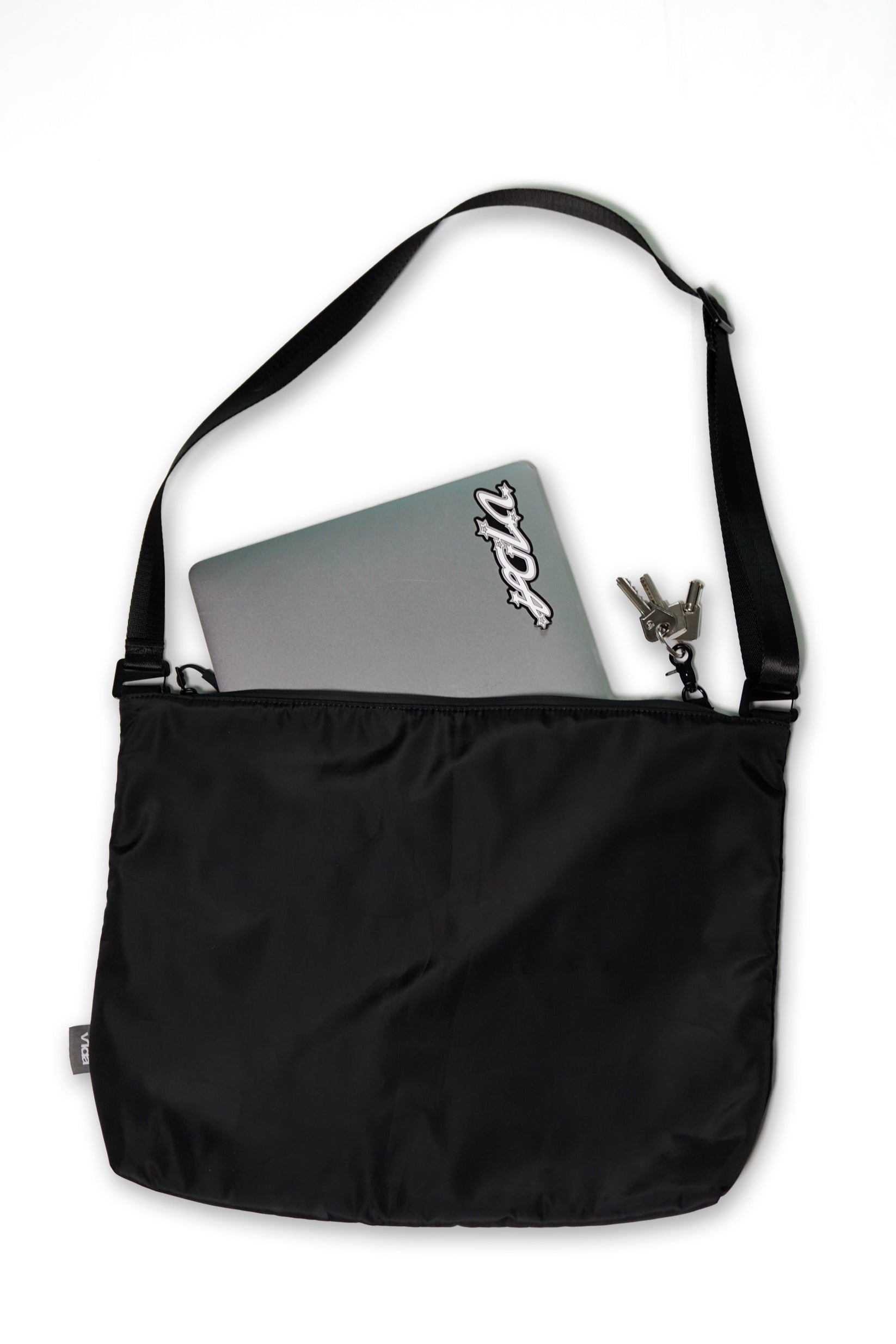 The V1da Everything Messenger Bag in black, shown on a white background, features a minimalist design with a partially visible laptop and keys tucked into one of its pockets.