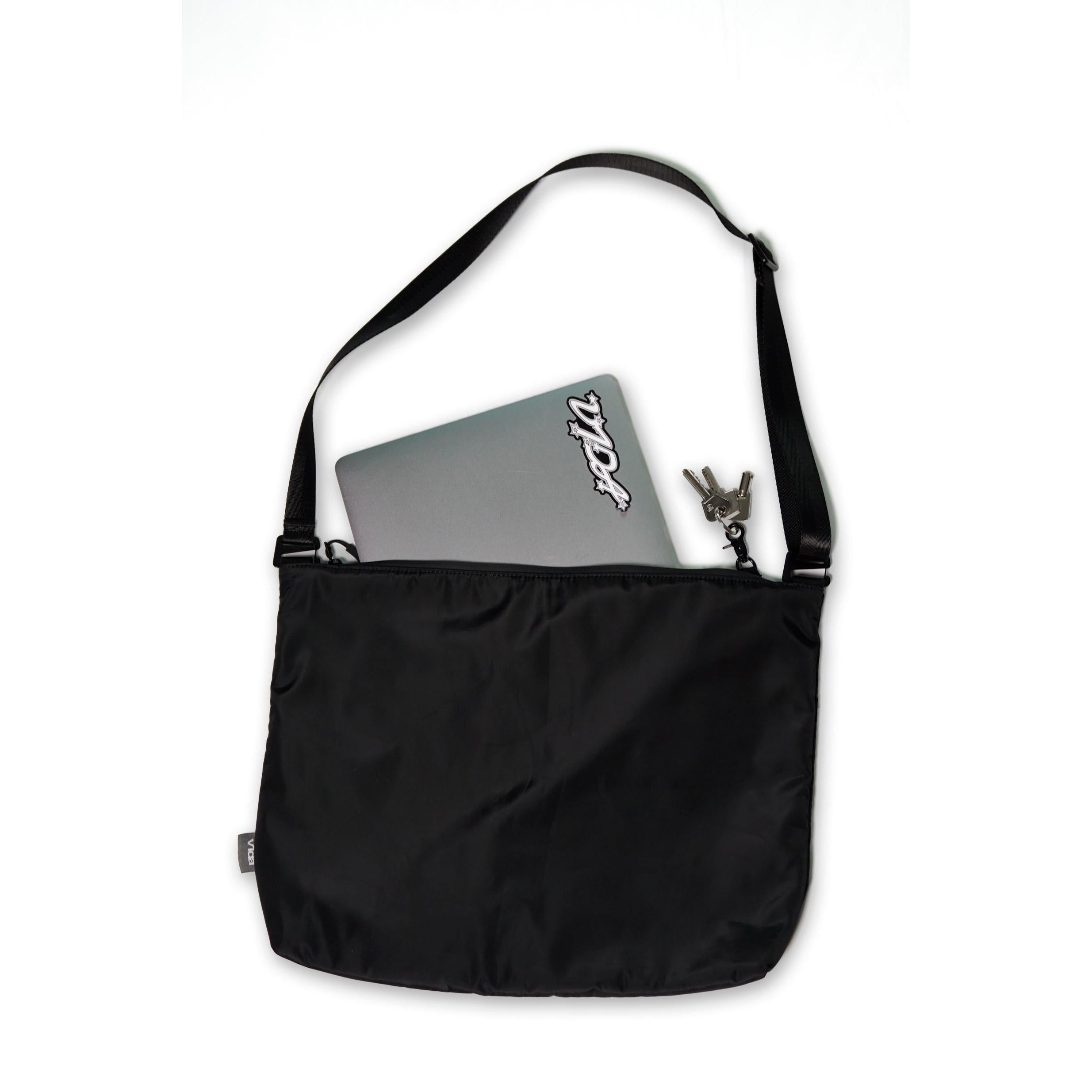 The V1da Everything Messenger Bag in black, shown on a white background, features a minimalist design with a partially visible laptop and keys tucked into one of its pockets.