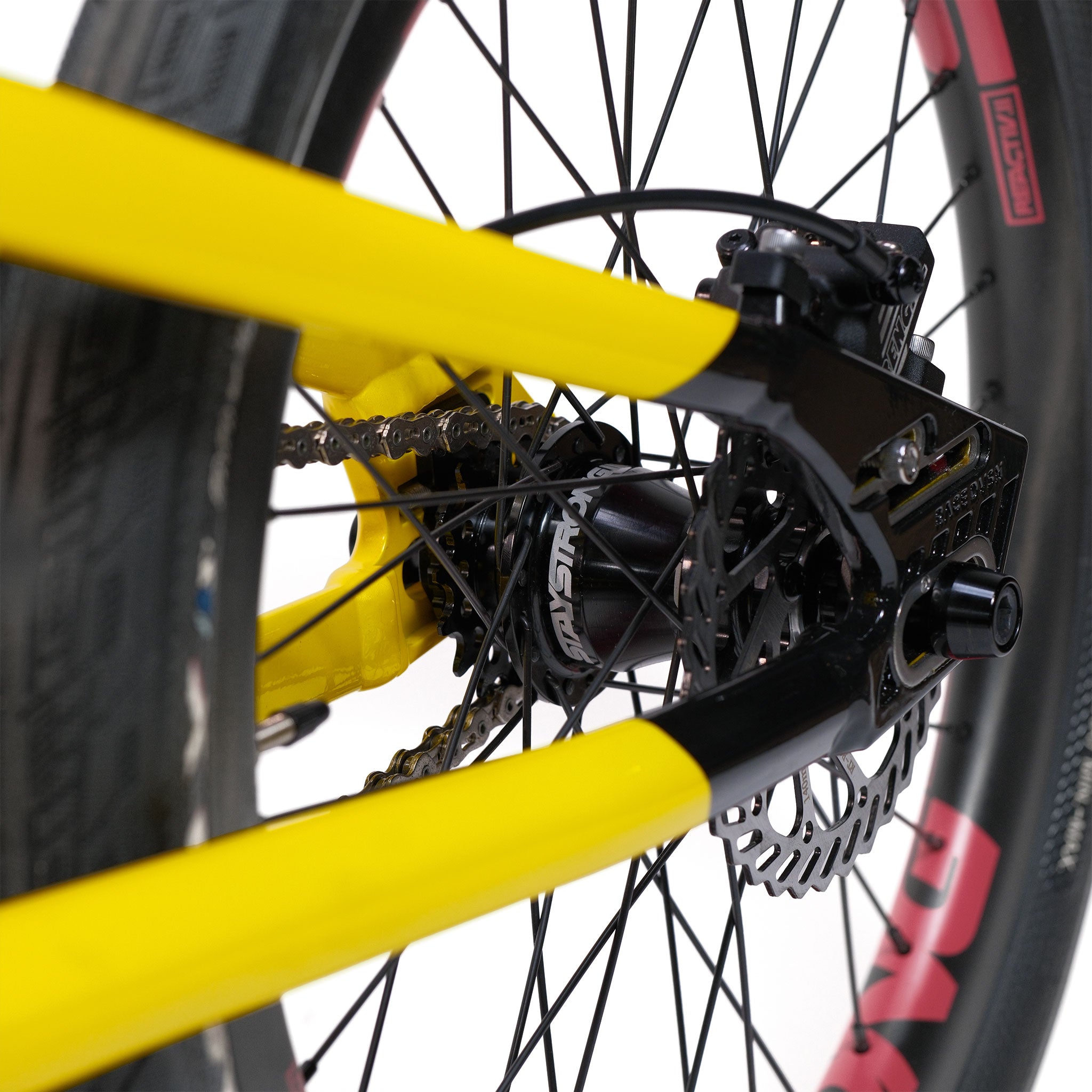 Close-up of the rear wheel and disc brake of a yellow Stay Strong V5.1 Pro XXXL Race Bike, featuring the chain, spokes, and branded hub—a striking detail often found on custom race bikes and premium aluminium frames.