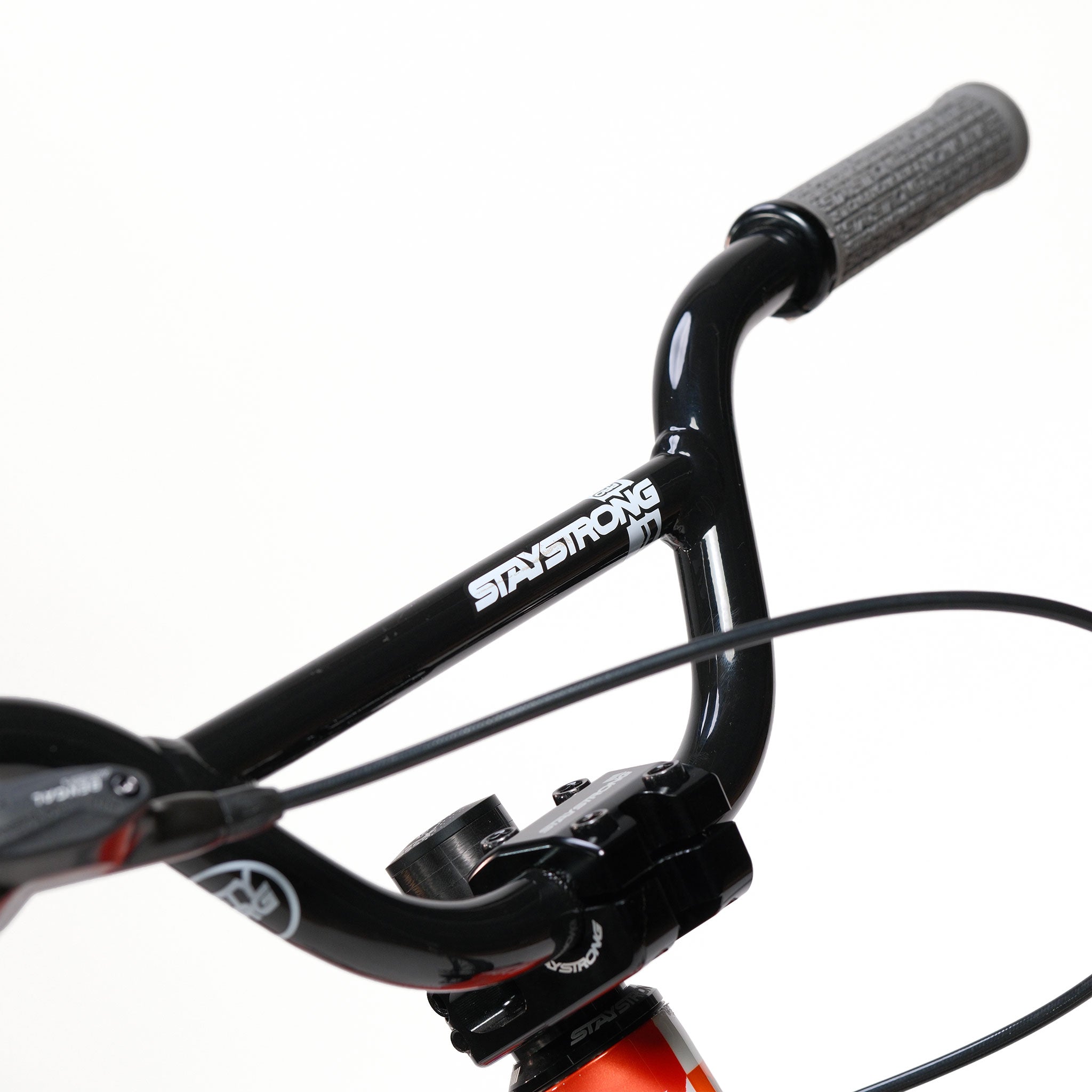 Close-up of the black handlebars of the Stay Strong V5.1 Junior Race Bike, featuring a "STAY STRONG" logo, visible handle grips, brake lever, and a section of the aluminium frame against a white background.
