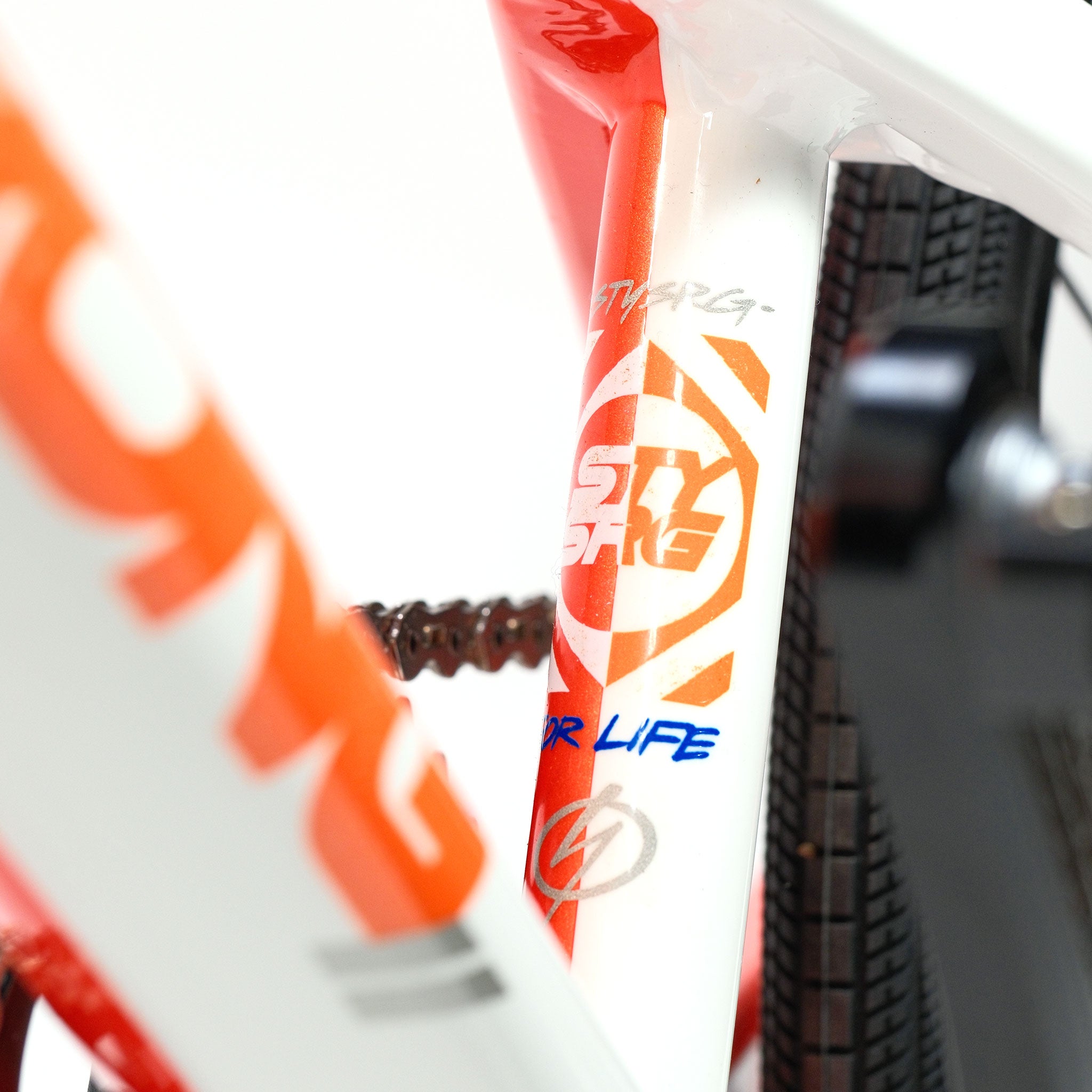 Close-up of a Stay Strong V5.1 Junior Race Bike, featuring an aluminium frame with orange and white graphics, partially displaying the text "STI BIKE" and "FOR LIFE," as well as showcasing the tire, chain, and disc brake in the background.