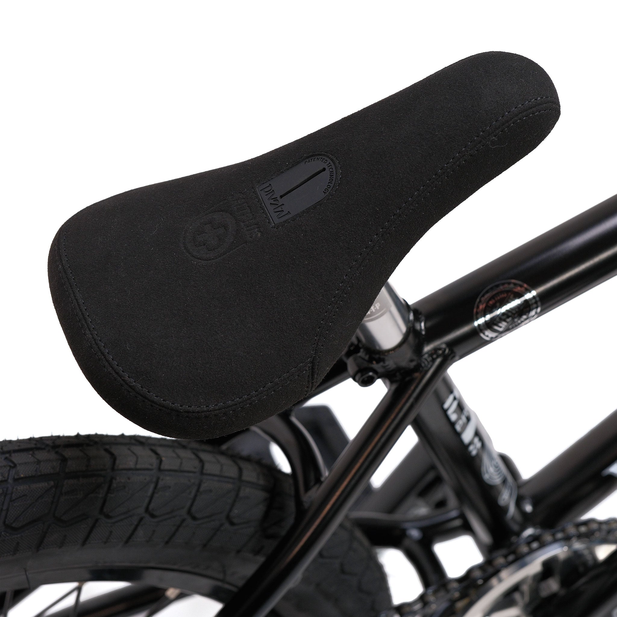 Close-up of a black padded bicycle seat attached to the Wethepeople Prodi-G-Unit 18 Inch Custom Bike’s black BMX frame, with part of the rear wheel and tire visible in the background. The setup features Eclat parts, highlighting attention to both style and performance.
