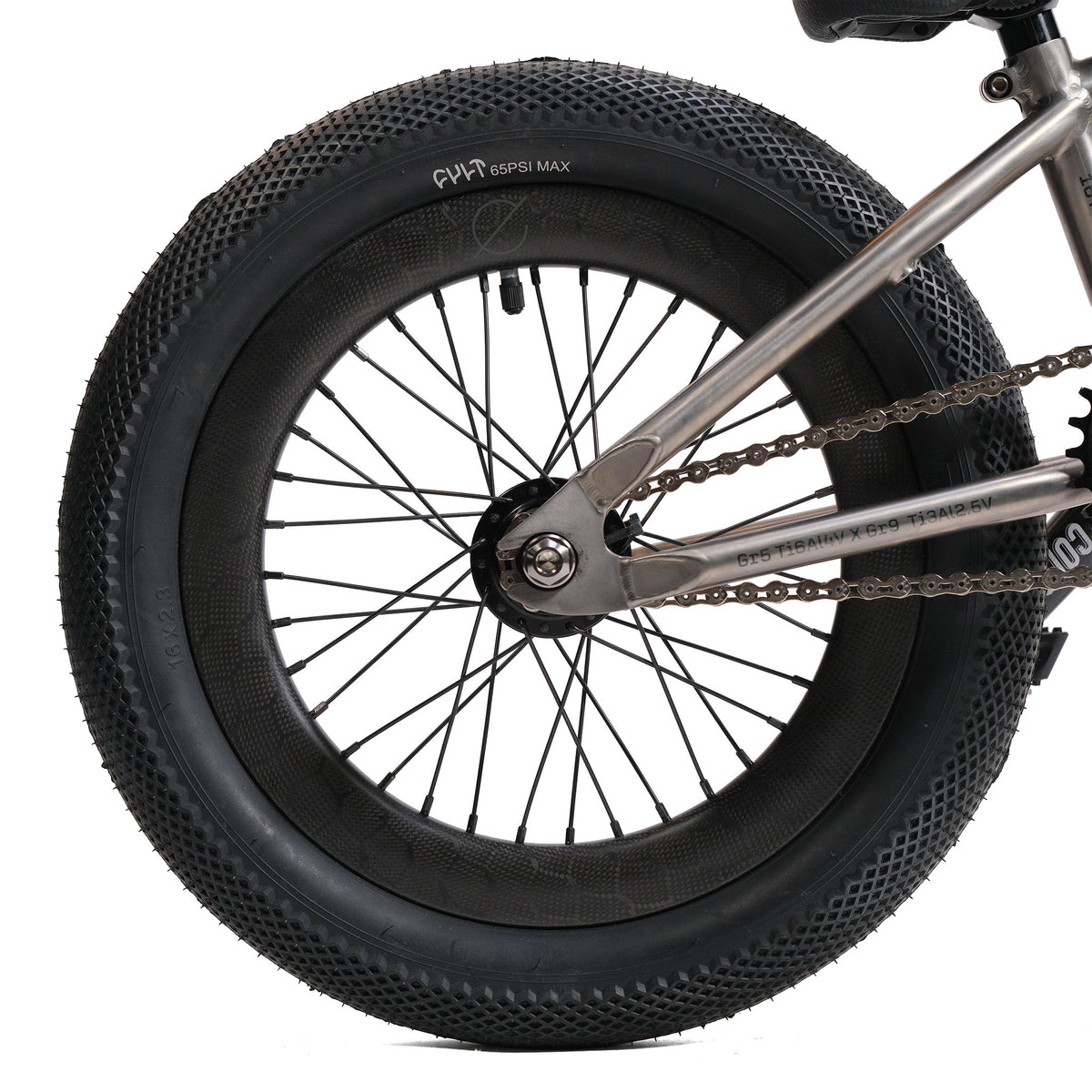 Close-up of the ION BMX Ti16 Custom Titanium 16 Inch Bike’s rear wheel, highlighting its thick tire tread, metal spokes, chain, and part of the titanium bike frame.