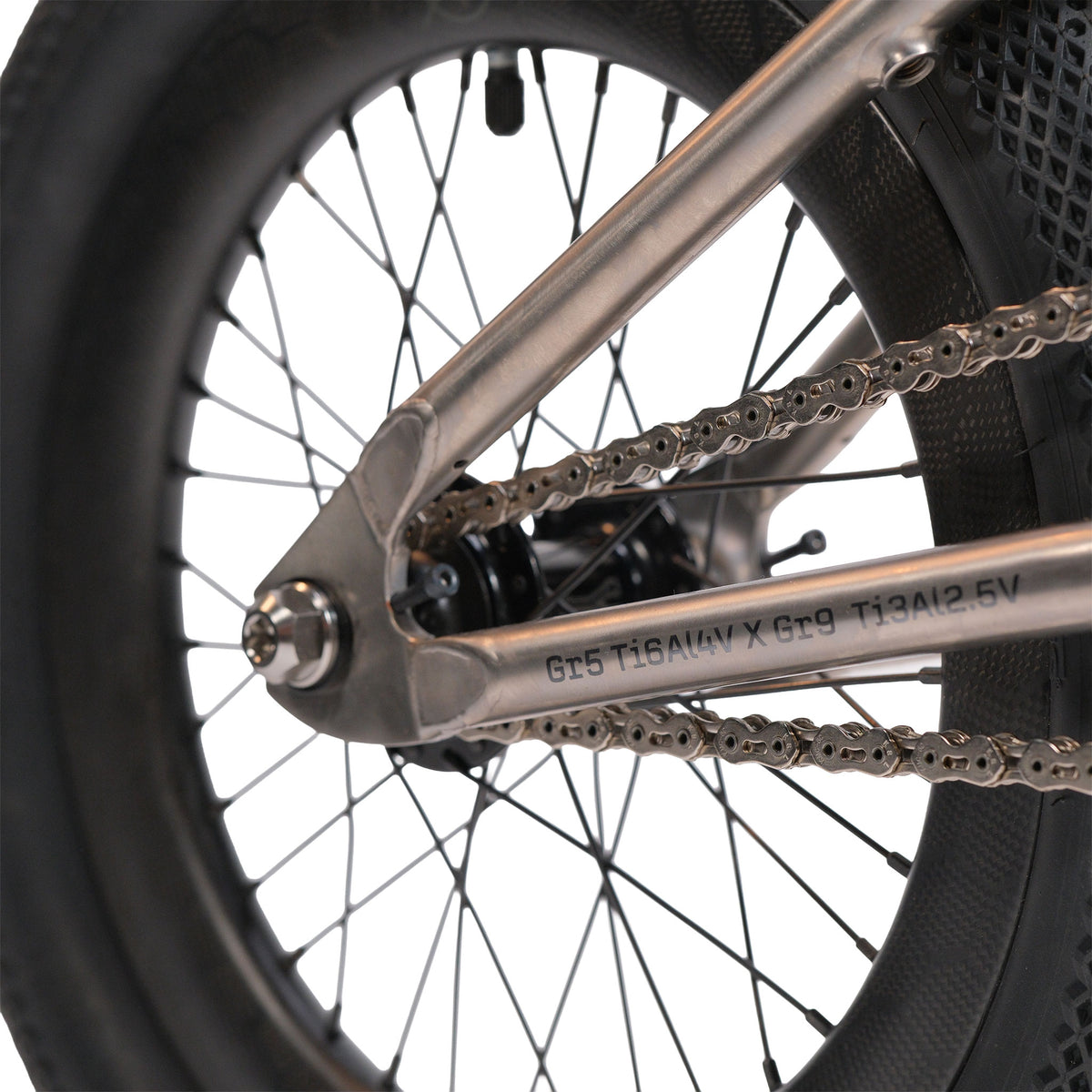Close-up of the ION BMX Ti16 Custom Titanium 16 Inch Bike’s rear wheel, highlighting the lightweight tire, metal spokes, chain, and a section of the titanium frame with engraved text.