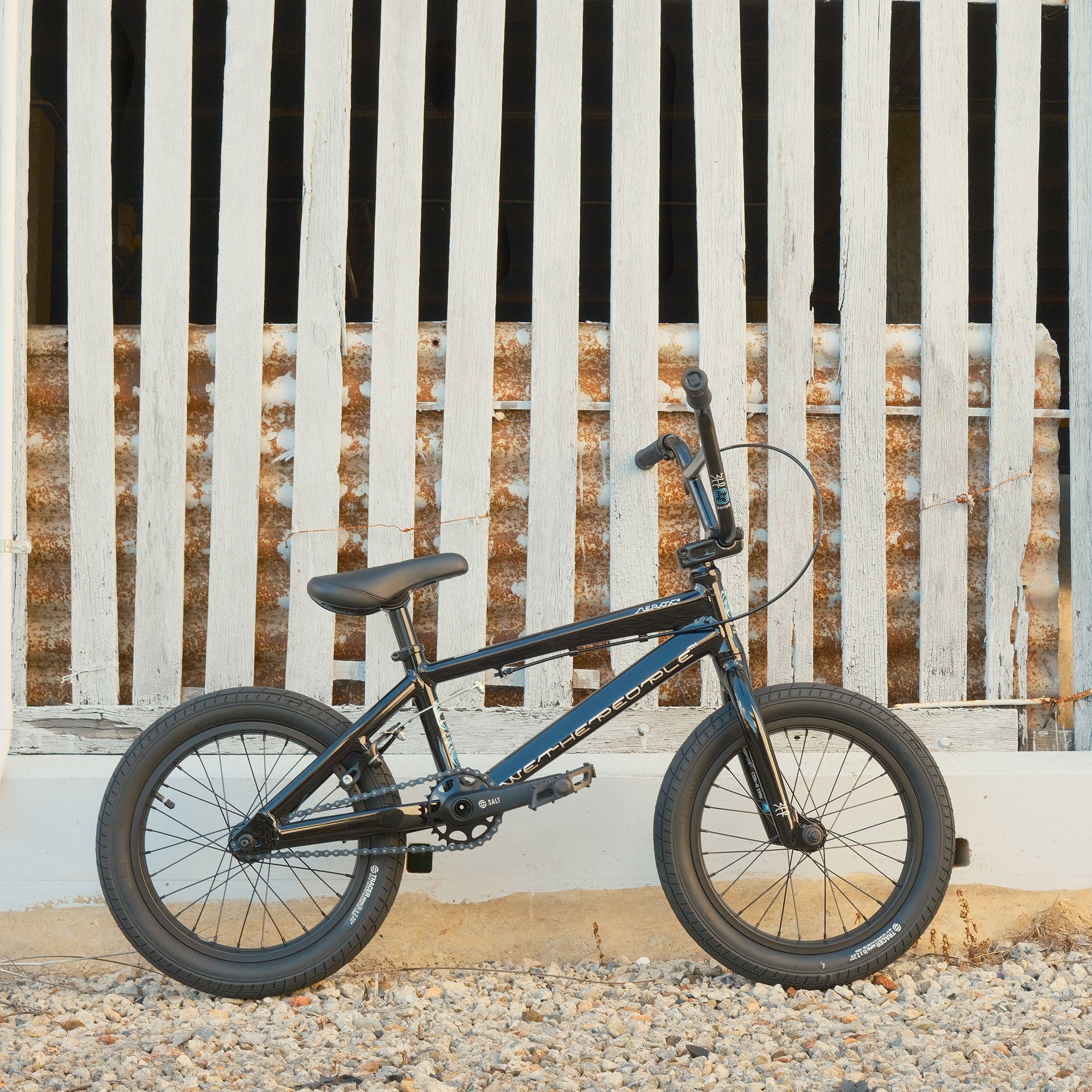A Wethepeople Aerox 16 Inch Bike, featuring a small black lightweight alloy frame, is parked on gravel in front of a white wooden fence with vertical slats.