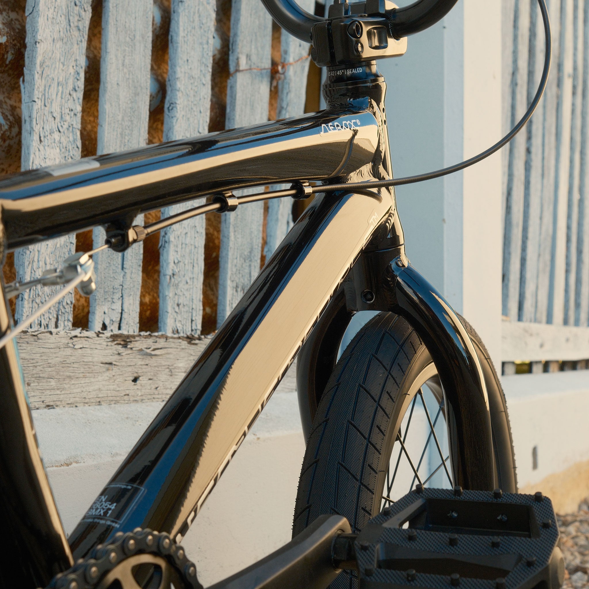 Close-up of the Wethepeople Aerox 16 Inch Bike’s black lightweight alloy frame and front wheel, set beside a white fence with a weathered blue wooden gate in the background.