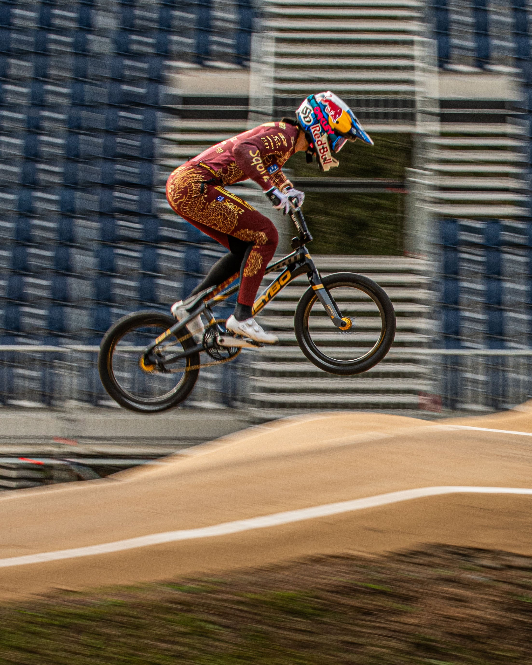 A BMX cyclist in maroon gear and a Red Bull helmet jumps over a dirt track, highlighting the Schwalbe SX-R BMX Tyre—the lightest race tyre—against blurred stadium seats.