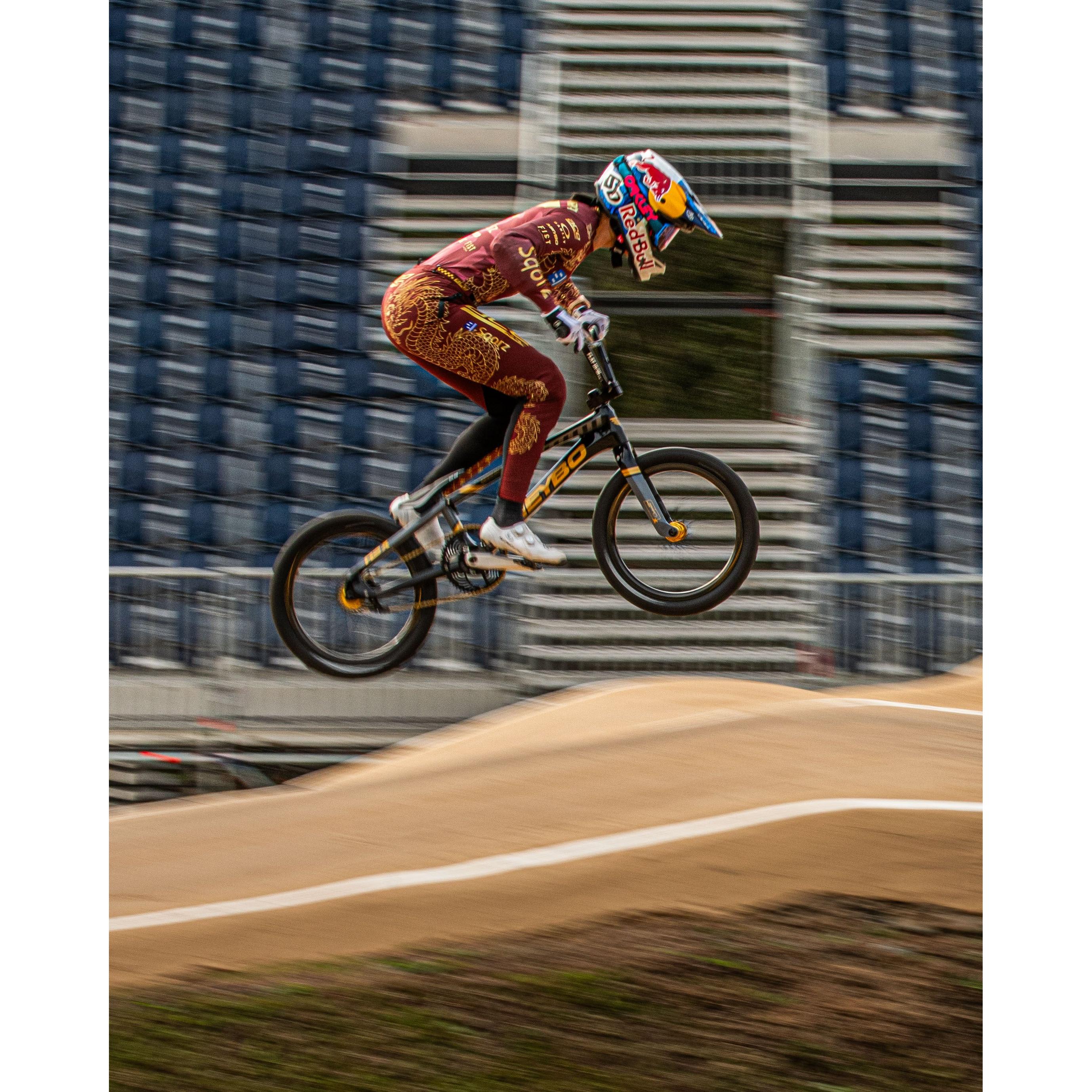 A BMX cyclist in maroon gear and a Red Bull helmet jumps over a dirt track, highlighting the Schwalbe SX-R BMX Tyre—the lightest race tyre—against blurred stadium seats.