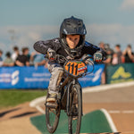 A young cyclist in race gear and helmet rides a BMX bike with number 111 at the National Championships track, captured in the LUXBMX Race Photo Package, with a blurred crowd visible in the background.