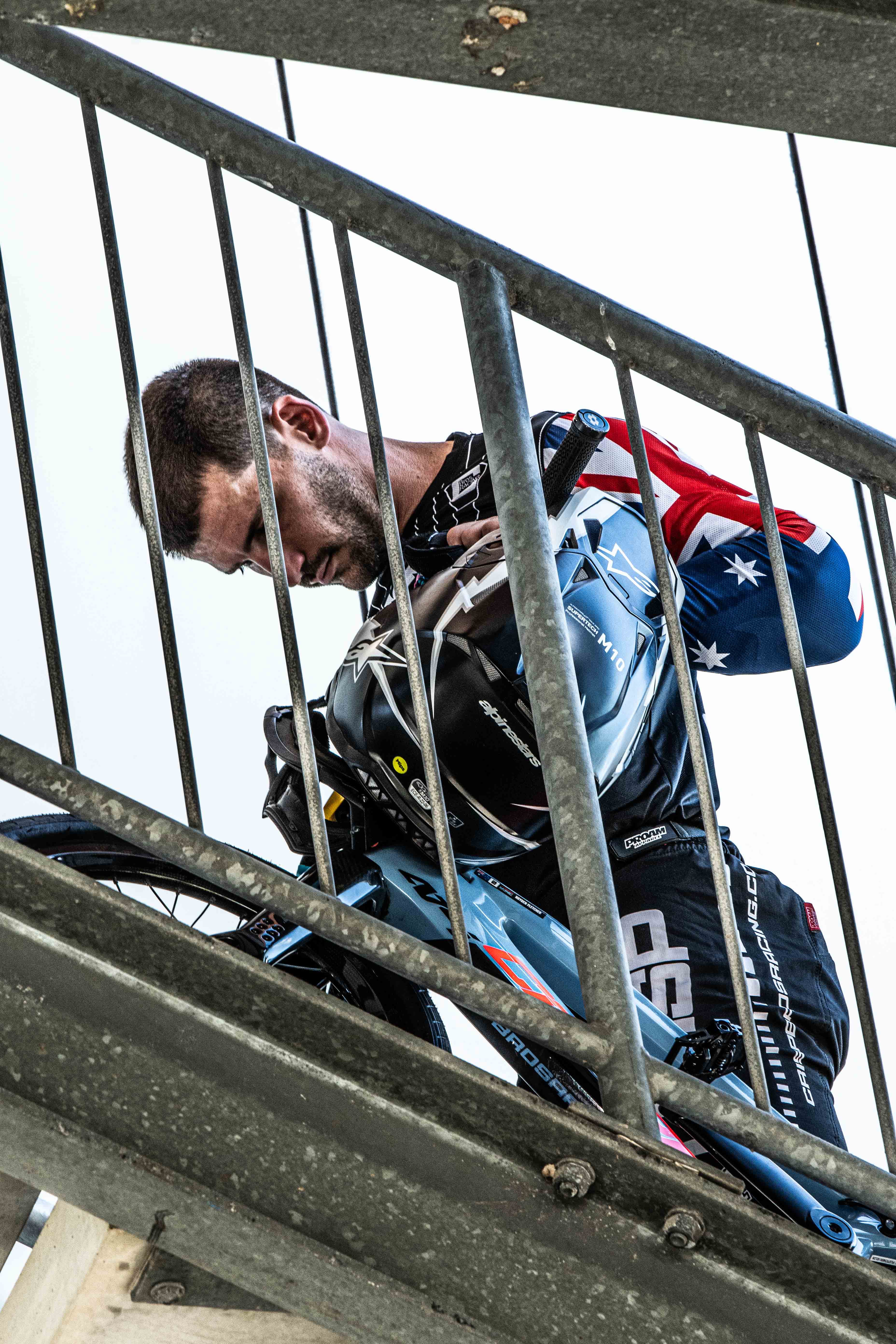 Captured during the National Championships, a man in motocross gear stands behind a metal staircase holding his helmet—partially obscured by railings. Photo from the LUXBMX Race Photo Package.