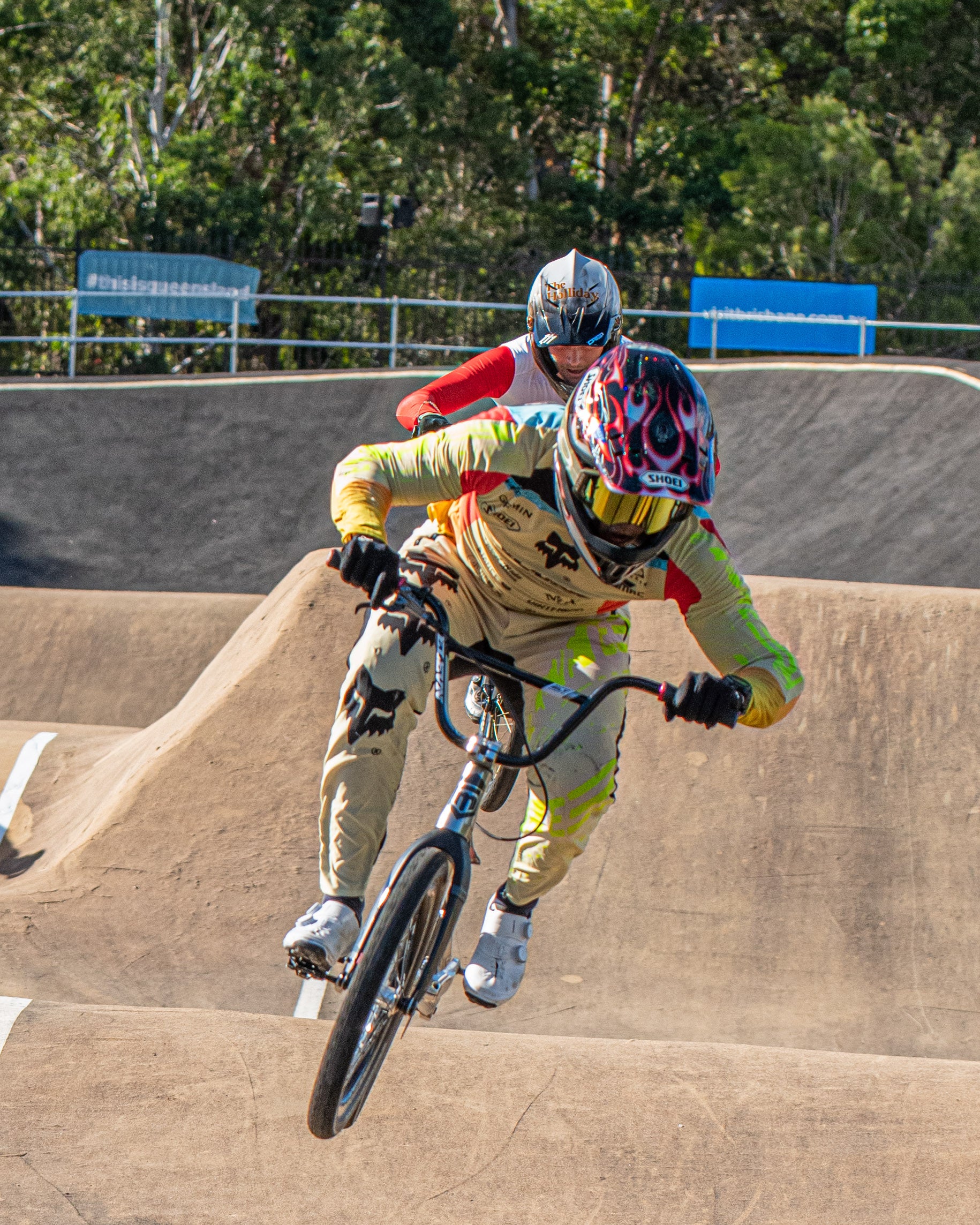 Capture the action at the world’s qualifier with the LUXBMX Race Photo Package: Two BMX cyclists in helmets and racing gear fly over a dirt jump, battling for a 2026 National Championships spot, with trees and barriers lining the course.