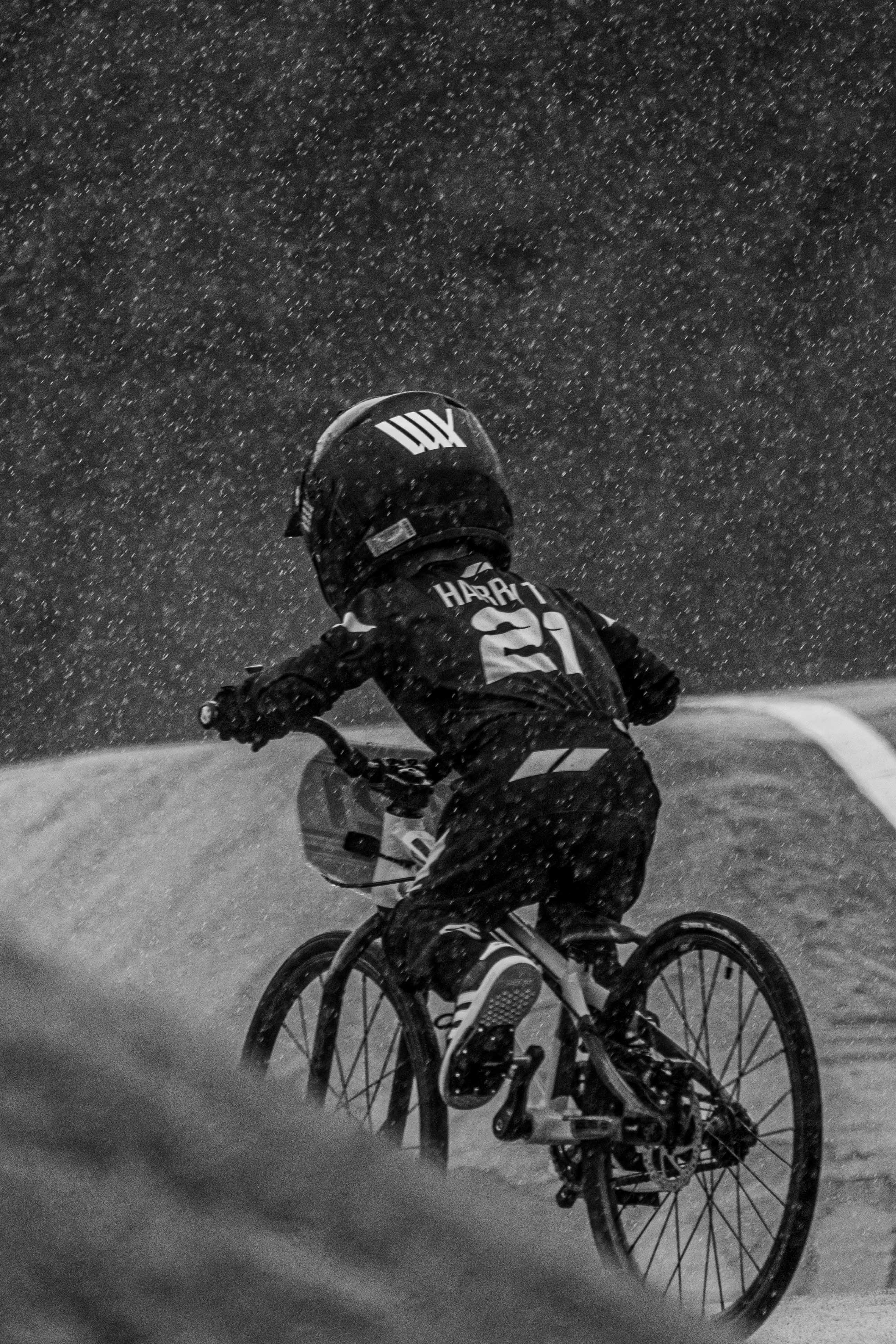 A cyclist in helmet and jersey rides a bike on a wet track in the rain during the National Championships, captured from behind for the LUXBMX Race Photo Package.