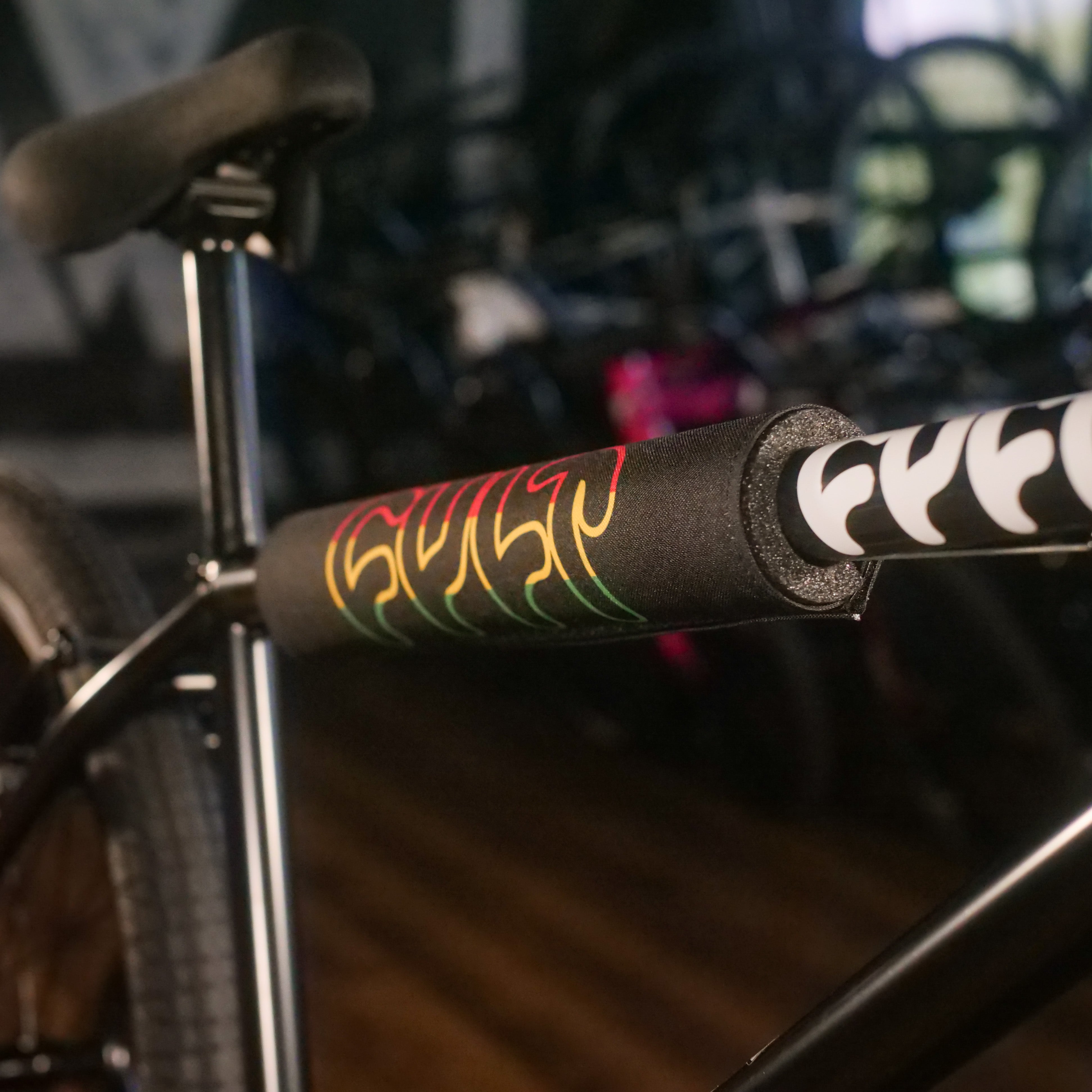 Close-up of the Cult Devotion 26 Inch Bike’s black Chromoly Frame top tube, fitted with a foam pad displaying vibrant flame graphics, captured in a bike shop setting.