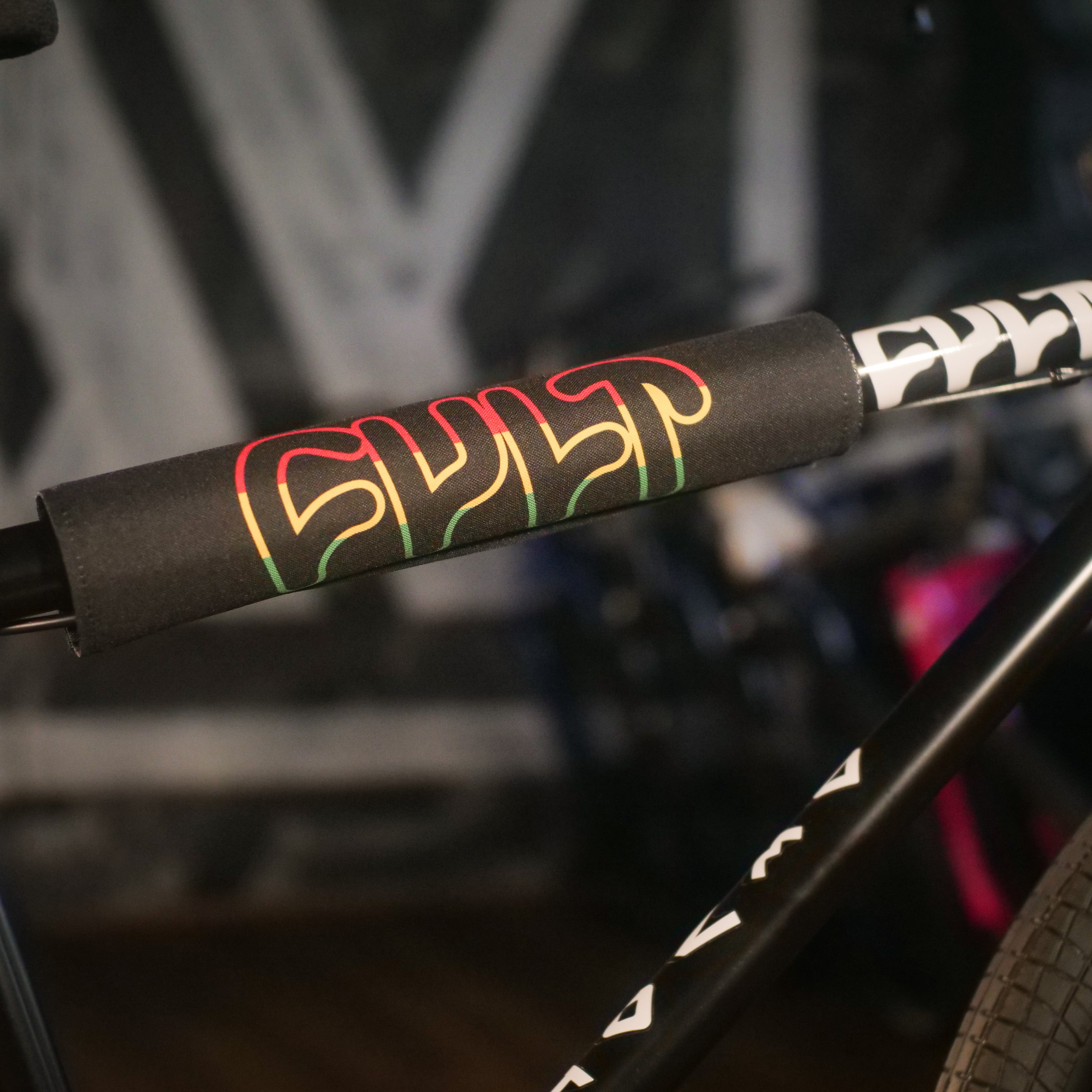 Close-up of a bike handlebar with a black foam pad displaying "CULT" in red, yellow, and green outlined letters. The blurred background suggests the sturdy Chromoly frame of the Cult Devotion 26 Inch Bike.