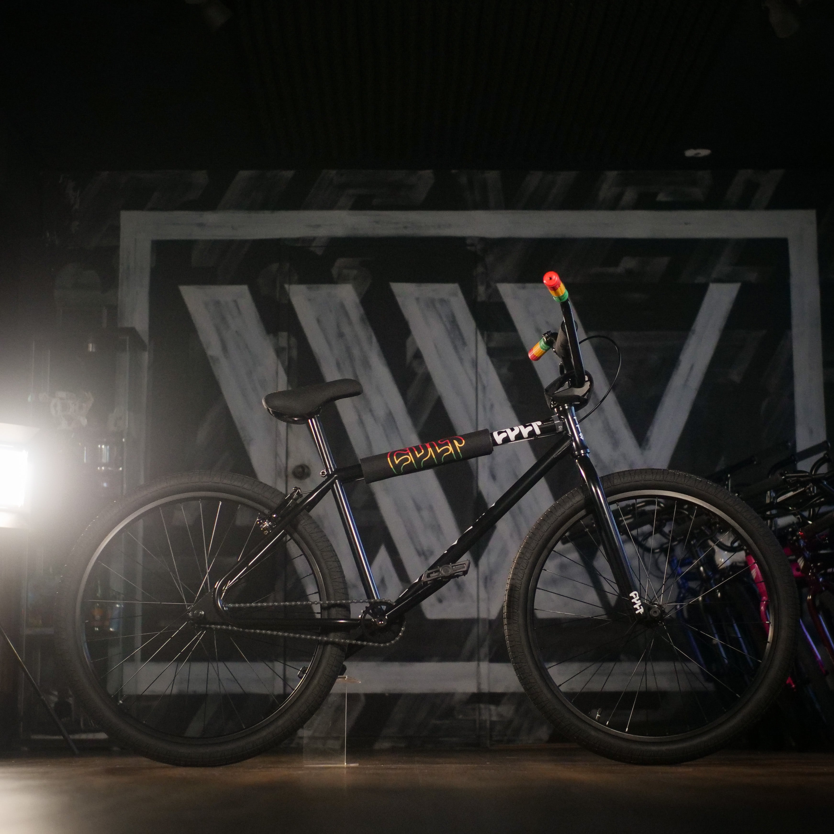 The Cult Devotion 26 Inch Bike, featuring a sturdy black Chromoly frame and colorful handlebar grips, is displayed indoors against a backdrop with large stylized white letters.