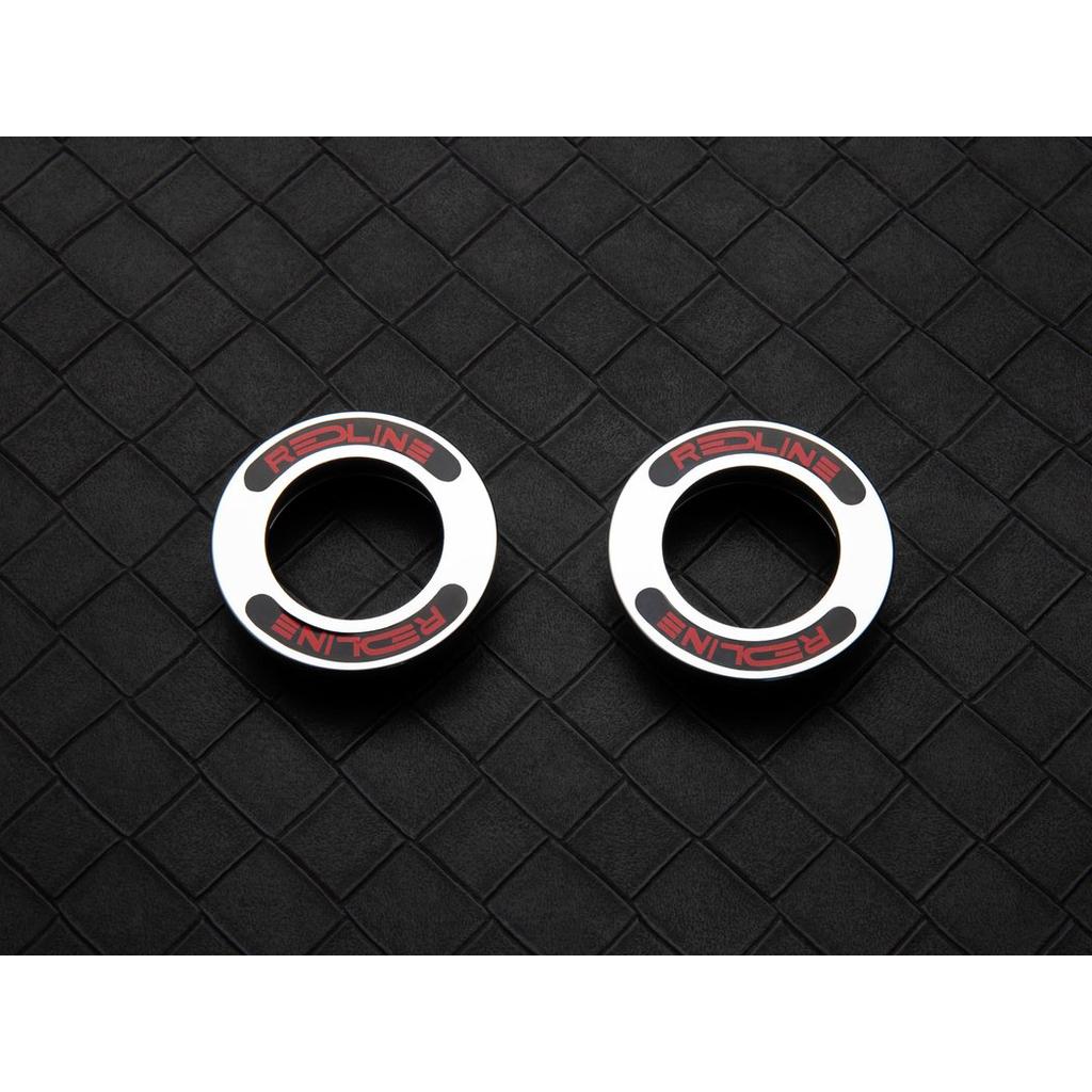 Two metallic circular objects labeled "REDLINE" are side by side on a textured black diamond-grid surface, resembling parts from the Redline Techmatic USA BB Set.