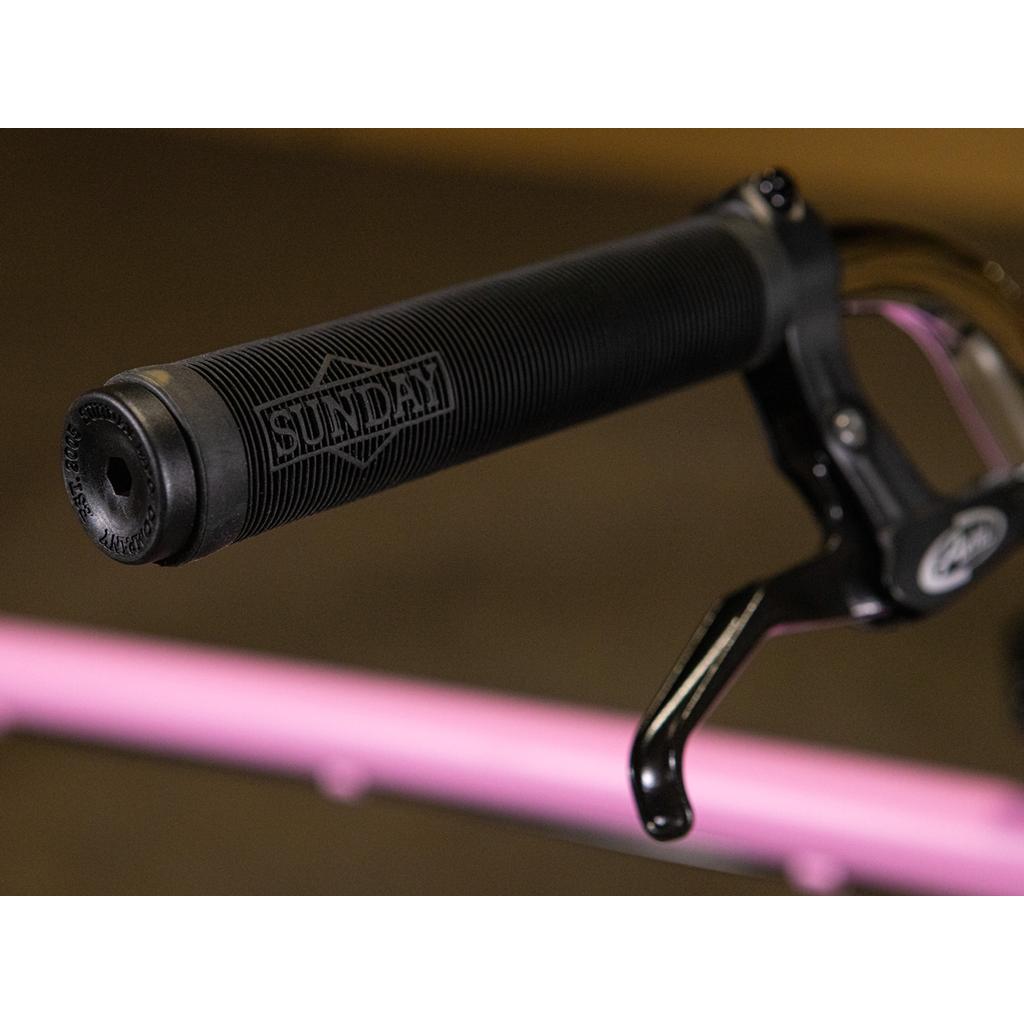 Close-up of a black handlebar grip labeled "SUNDAY" beside a brake lever, with a blurred pink horizontal bar in the background—a signature element of the Sunday High C 29 Inch Bike, renowned for its durable chromoly frame and smooth-riding 29" cruiser design.