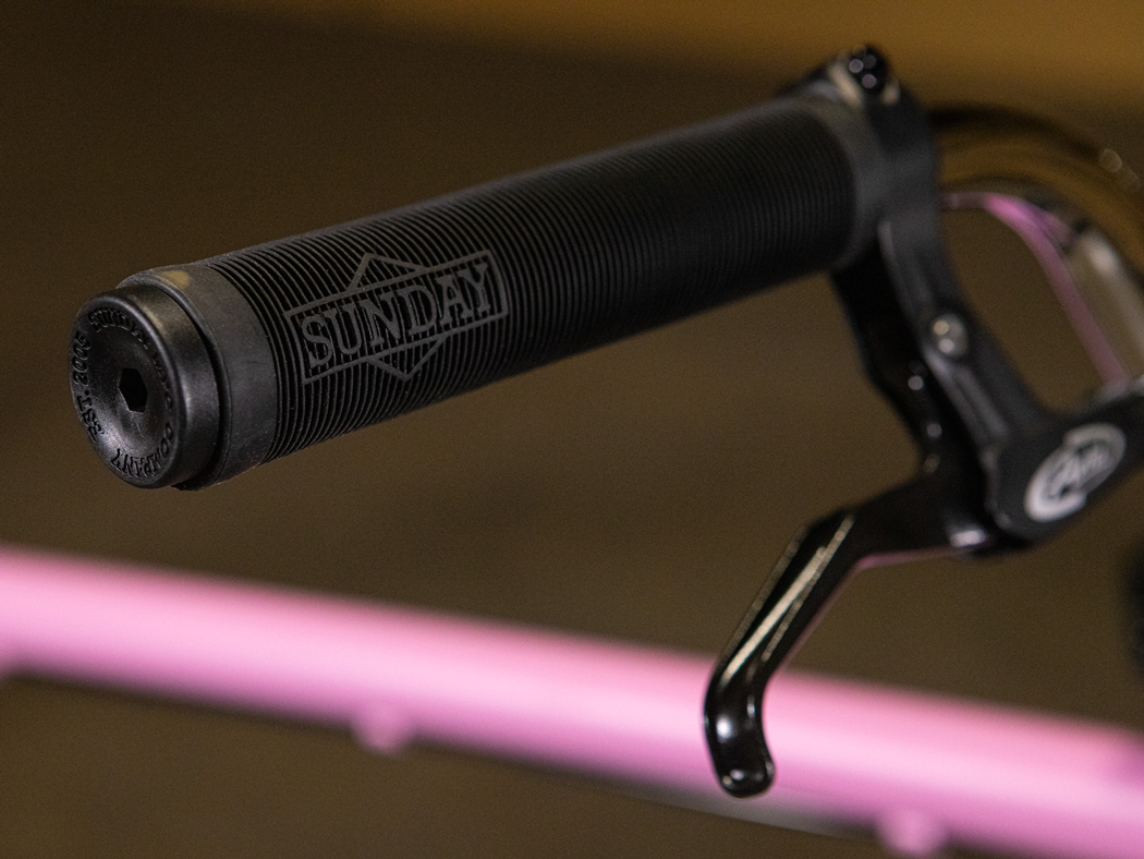 Close-up of a black handlebar grip labeled "SUNDAY" beside a brake lever, with a blurred pink horizontal bar in the background—a signature element of the Sunday High C 29 Inch Bike, renowned for its durable chromoly frame and smooth-riding 29" cruiser design.