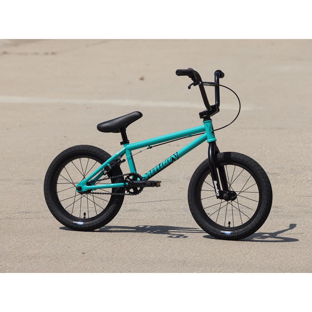 The Sunday Primer 16 Inch Bike (2025), featuring a teal frame, black seat, black handlebars, and thick tires with Pro-Style Geometry, is parked on a concrete surface.