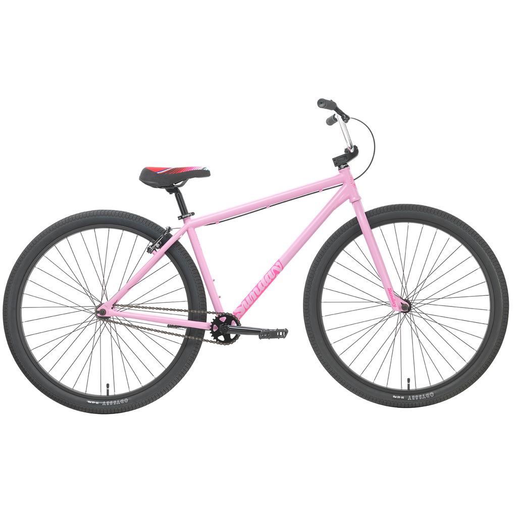 The Sunday High C 29 Inch Bike is pictured in pink with black tires, black handlebars, a red and black seat, and a simple single-speed design without visible gears or accessories. Shown from the side on a white background, this chromoly cruiser offers both style and reliability with its durable Odyssey components.