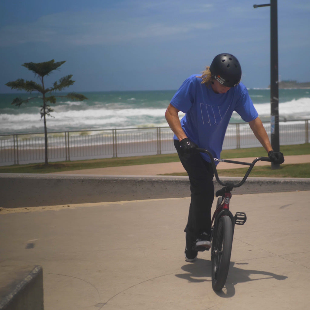 A man riding a LUXBMX bike on a sidewalk in front of the ocean, showcasing the LUXBMX Eclipse Tee - Violet.