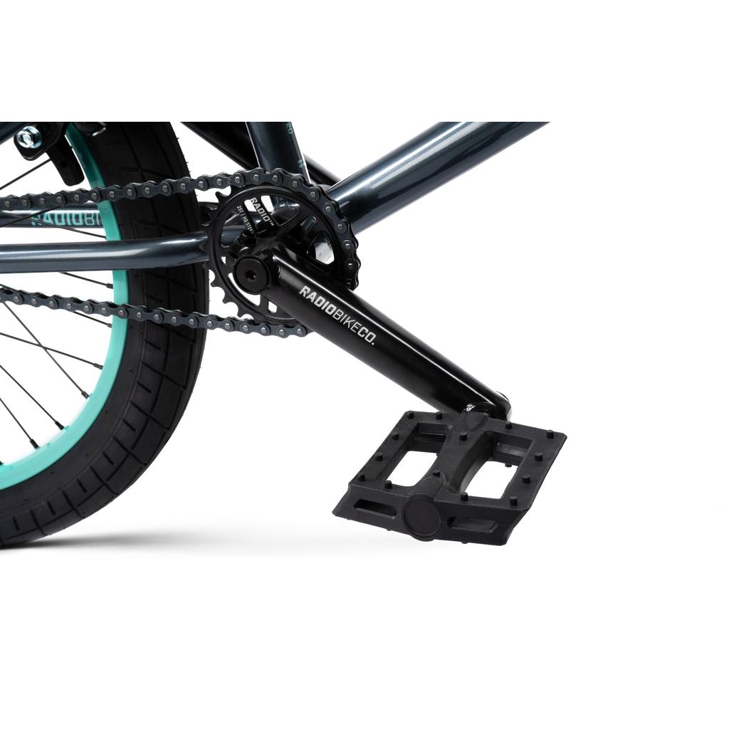 Close-up of the Radio Saiko 20 Inch Bike crankset, chain, and pedal attached to a metallic frame with a black tire featuring a teal Salt Tracer Tyres sidewall.