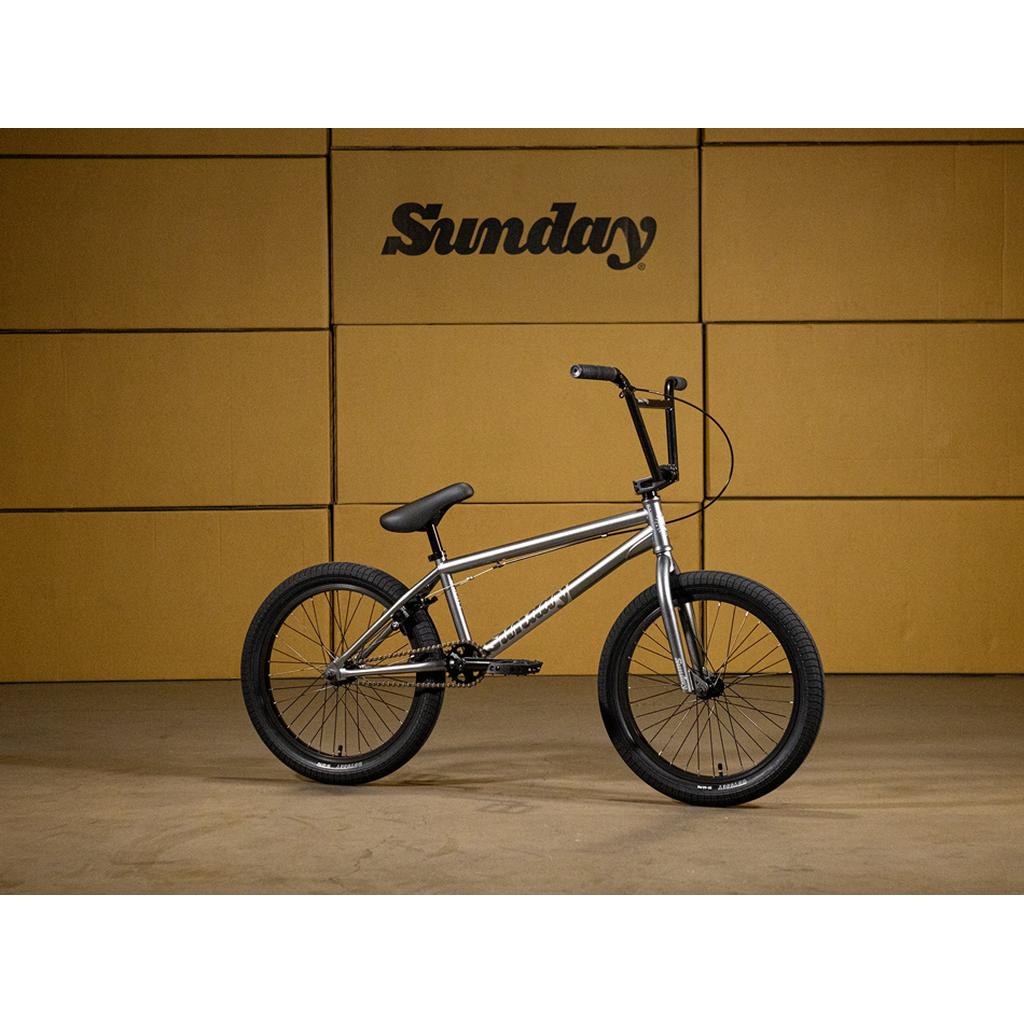 The Sunday Scout 20 Inch Bike, finished in silver, is showcased indoors against a yellow wall with the "Sunday" logo in bold black text. This BMX bike boasts high-quality Odyssey components, offering a perfect blend of style and performance.